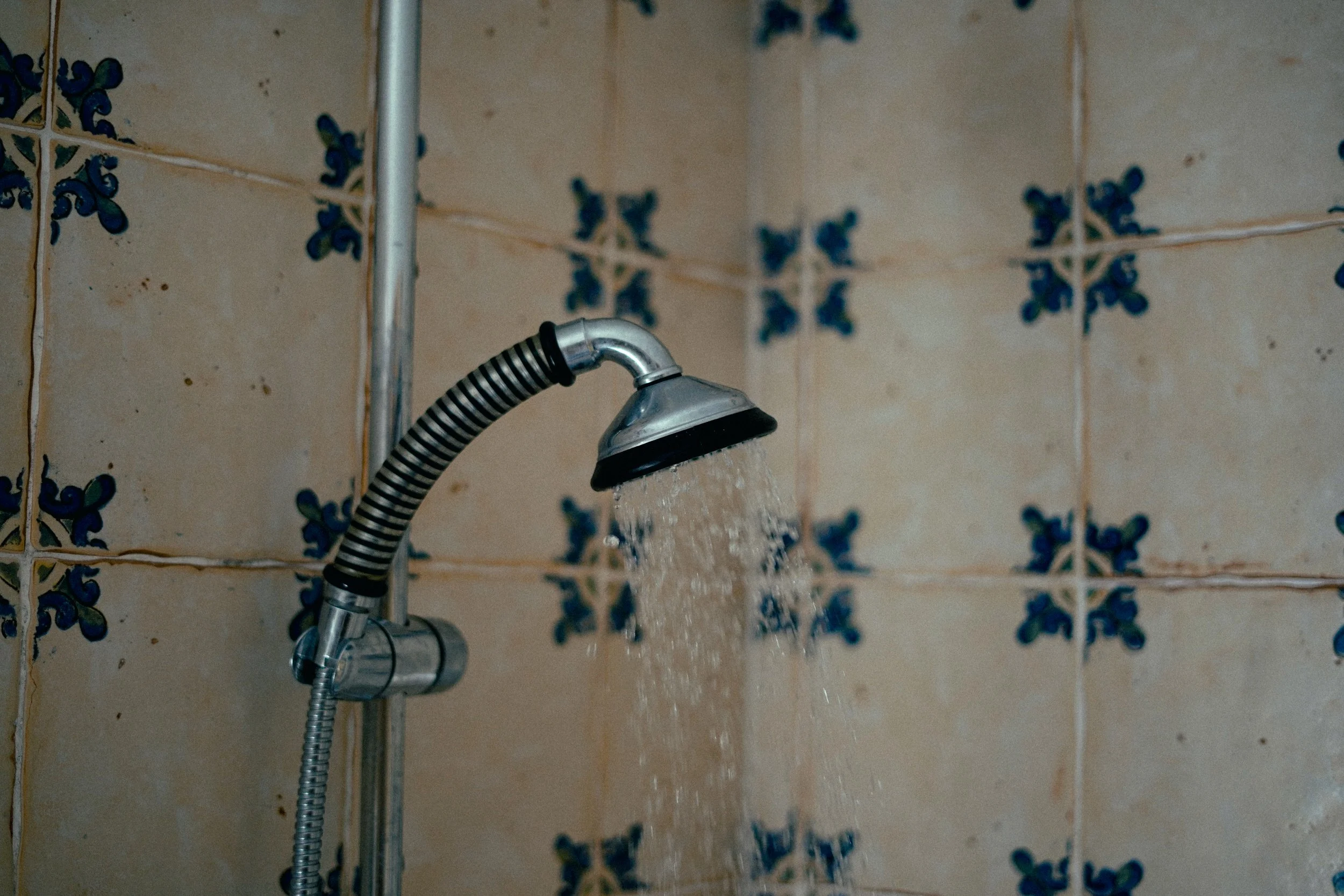 Ein Duschkopf, aus dem Wasser fließt, an einer Wand mit beige Kacheln mit blauen Mustern.