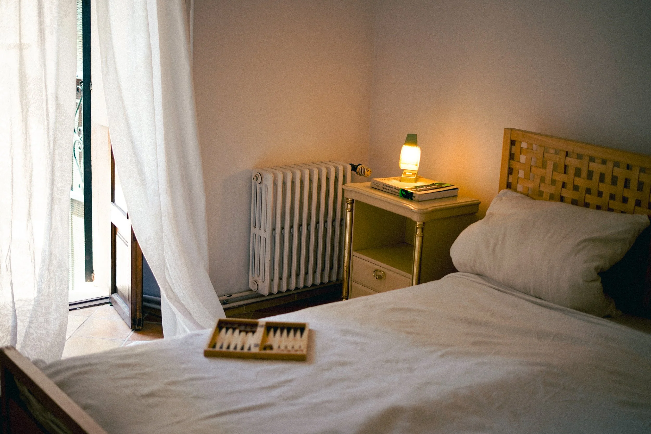 Ein Schlafzimmer mit einem Bett, Nachttisch mit Büchern und einer Lampe, Gardinen vor einem Balkonfenster.