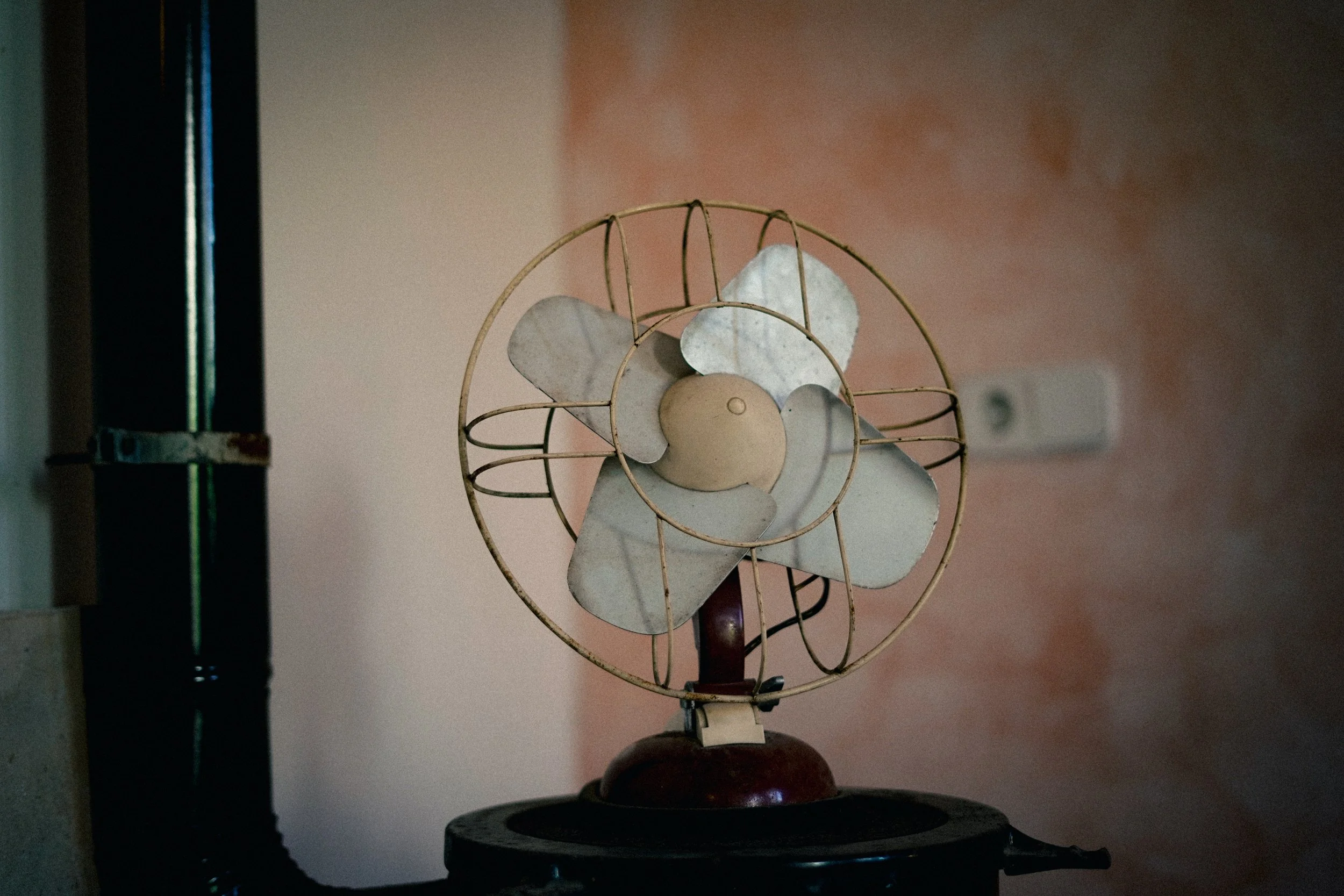 Alter elektrischer Ventilator mit Flügeln, auf einem dunklen Tisch, vor einer Wand mit Steckdose, in einer unbelebten Umgebung.