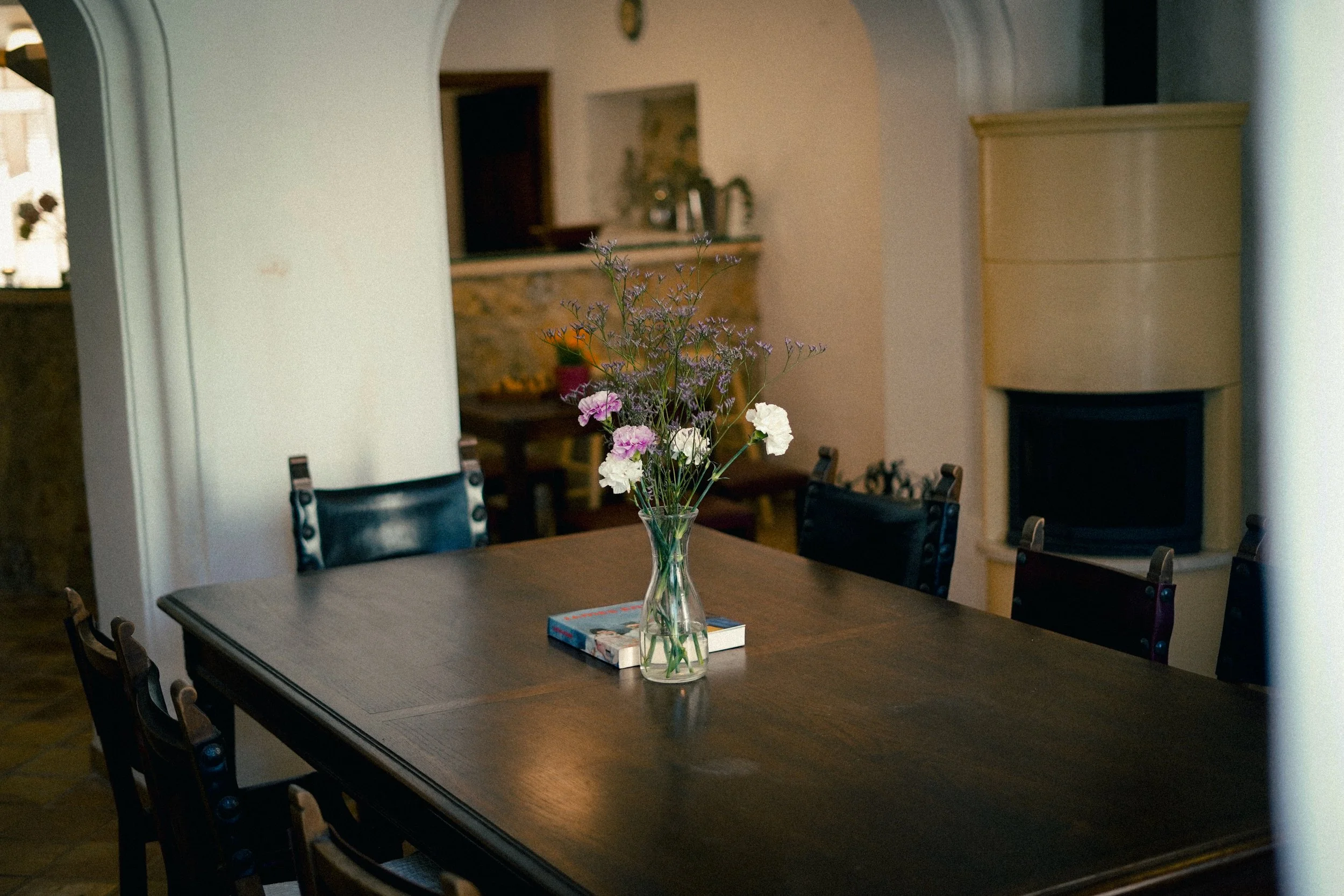 Ein Esstisch mit einer Vase voller Blumen in der Mitte. Hintergrund zeigt eine offene Küche mit Holzregalen und Kamin.