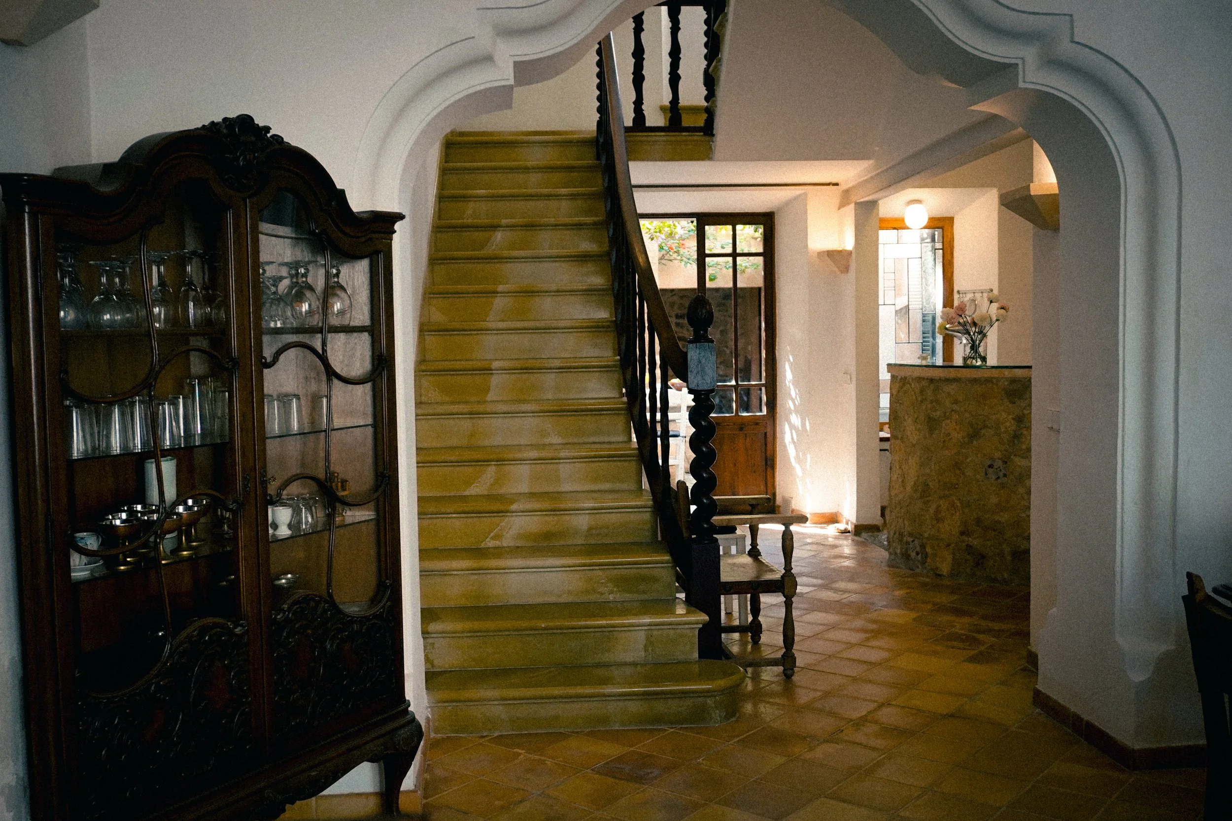 Innenaufnahme eines Flurs mit Treppe, einer großen Vitrine mit Gläsern, einem Fenster im Hintergrund, einer eingehenden Tür, einer Vase mit Blumen auf einer Theke und Kacheln auf dem Boden.