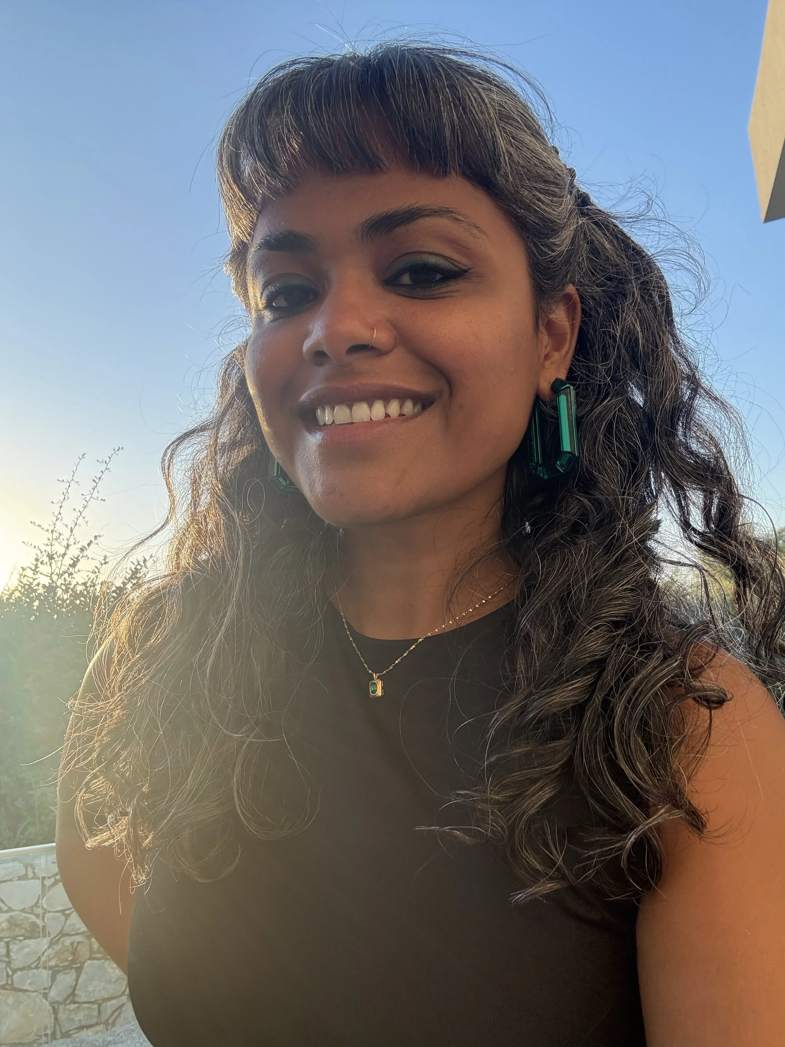 Close-up of a smiling woman with long curly hair, wearing a black top, green earrings, and a gold necklace, outdoors with a clear blue sky in the background.