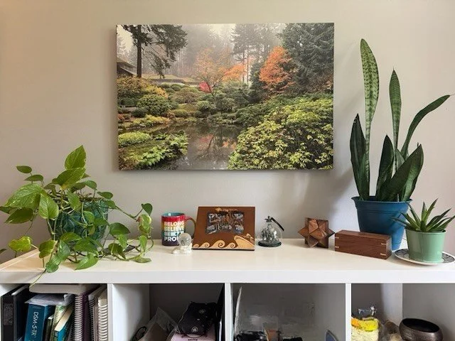 A white shelf with various potted plants, decorative objects, and a framed photo. A large landscape art piece hangs on the wall behind the shelf.