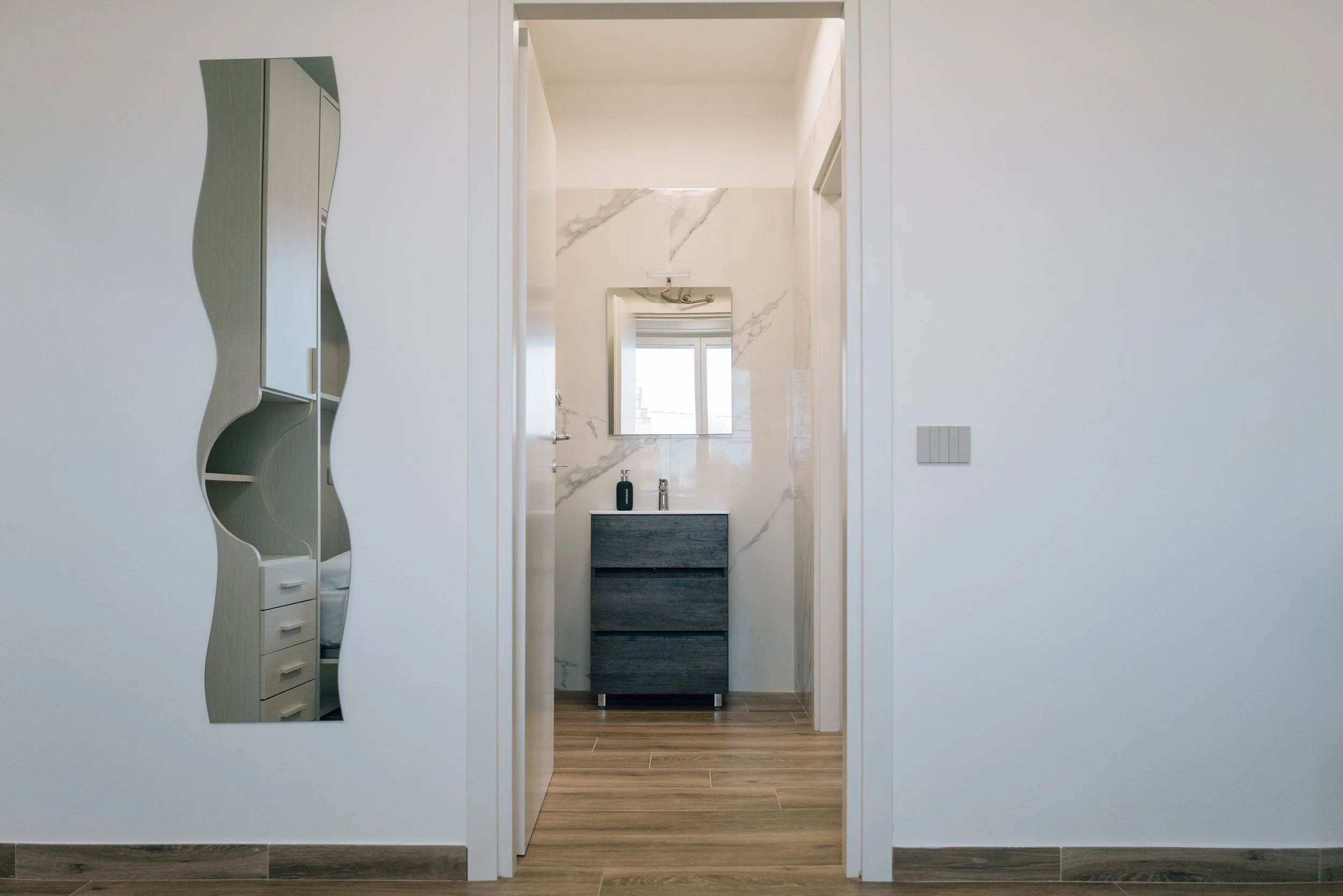 Interno di un appartamento con pareti bianche, uno specchio curvilineo sulla parete a sinistra, un mobiletto grigio scuro con specchio sopra nel bagno che si vede attraverso una porta aperta, piastrelle di marmo chiaro sul muro e pavimento in legno.
