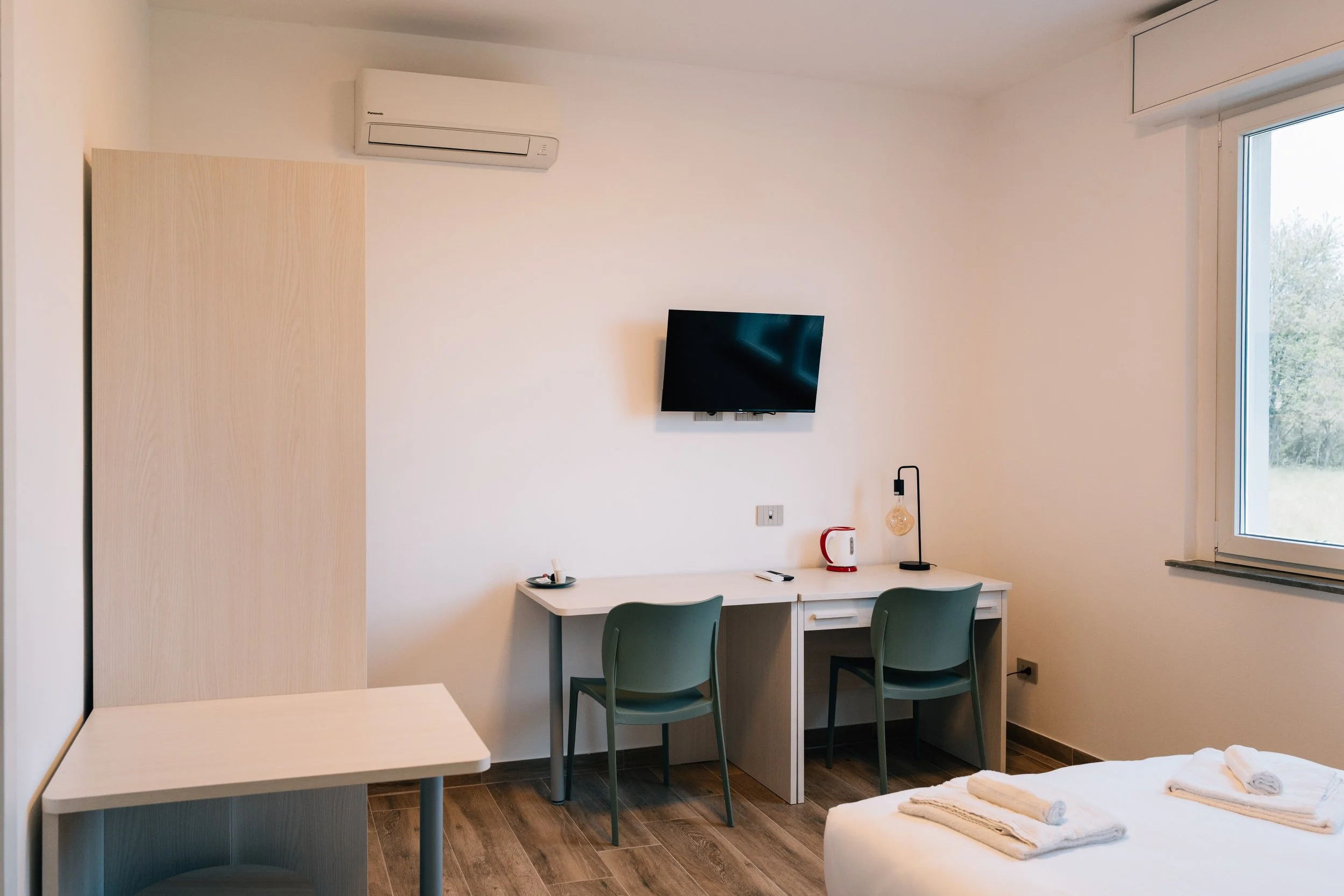 Interno di camera d'hotel con letto, scrivania, sedie, televisore, lampada, aria condizionata, finestra con vista su natura.