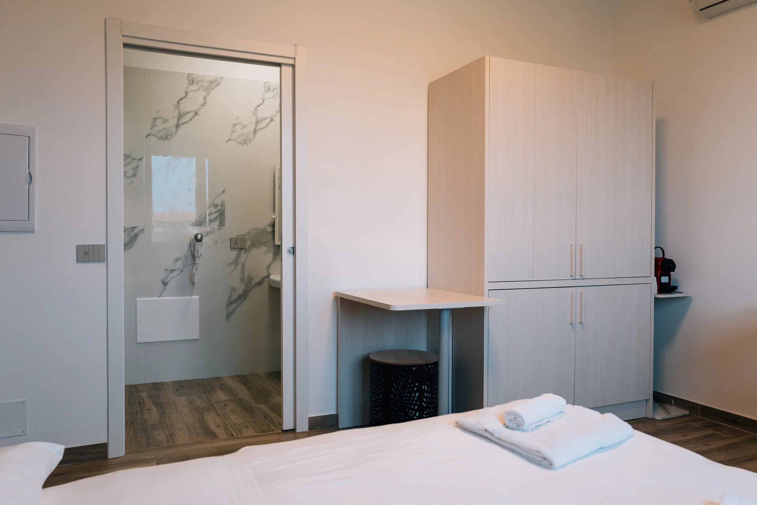 Stanza con armadio e scrivania, vista bagno con pareti in marmo, letto con asciugamani e roullette di carta, sulla destra macchina da caffè.