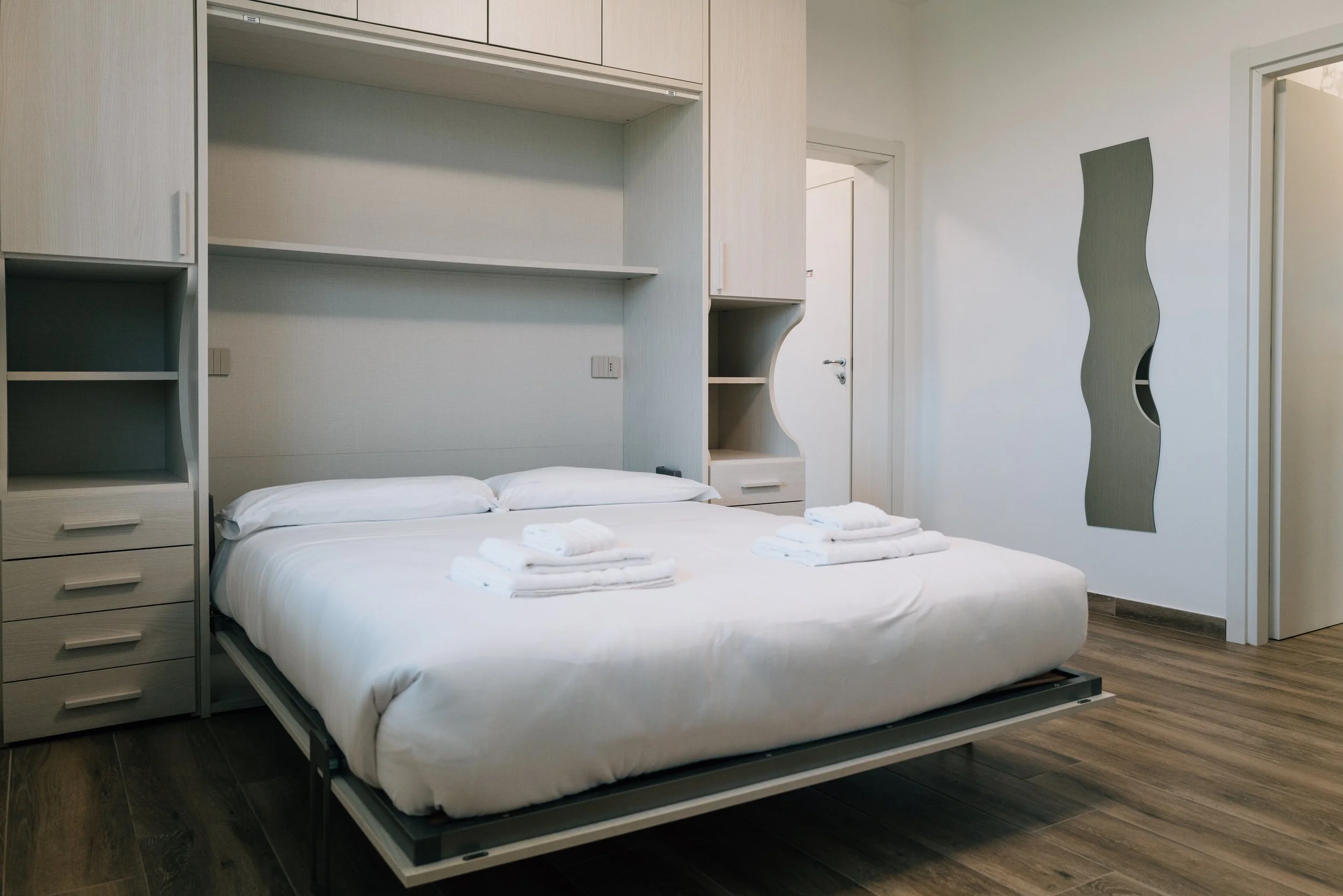 Stanza d'albergo con letto matrimoniale e asciugamani piegati sul letto, pareti bianche e arredi moderni in legno chiaro.