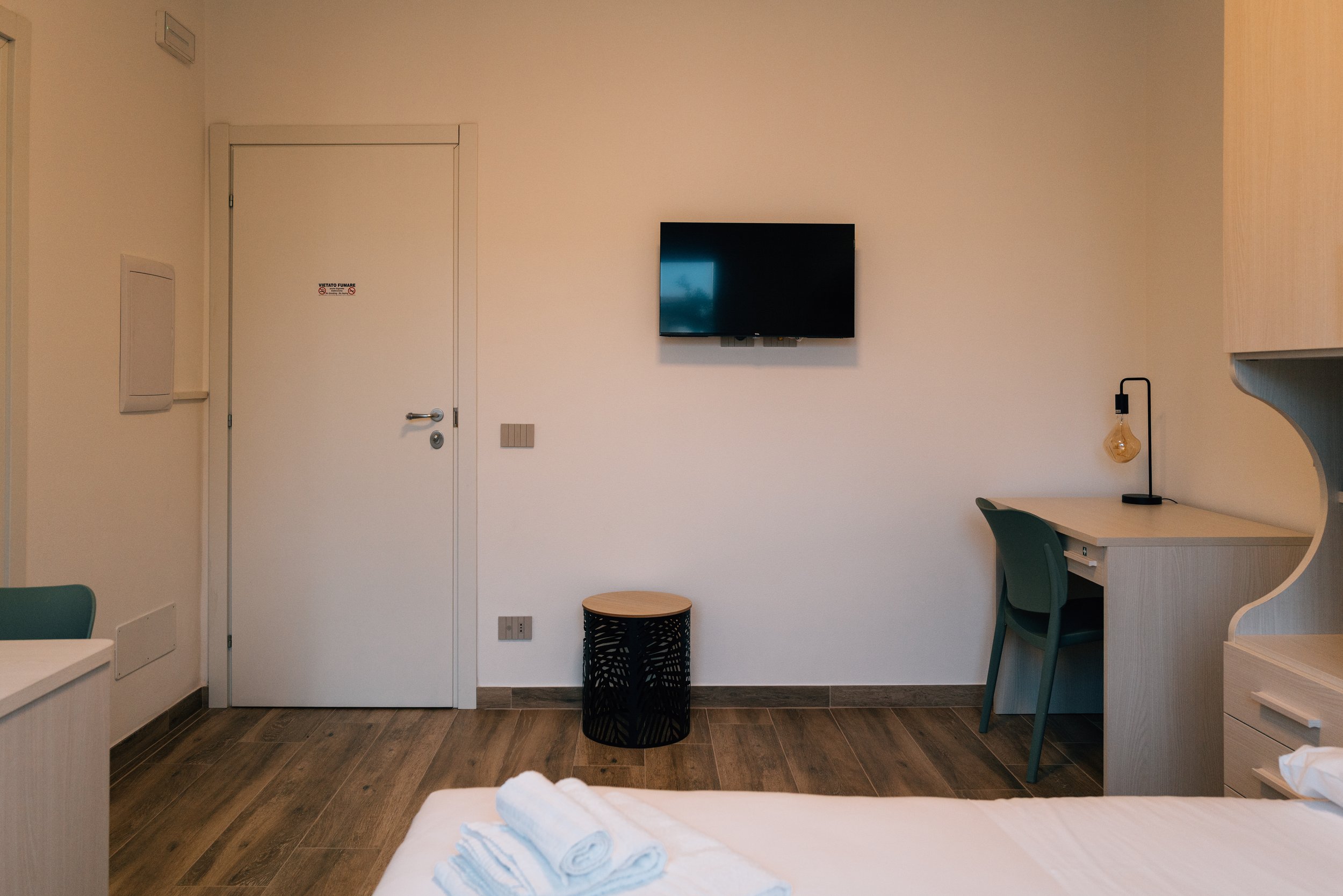 Camera da letto con pareti bianche, pavimento in legno, TV montata sulla parete, scrivania con lampada, sedia, e letto con asciugamani.