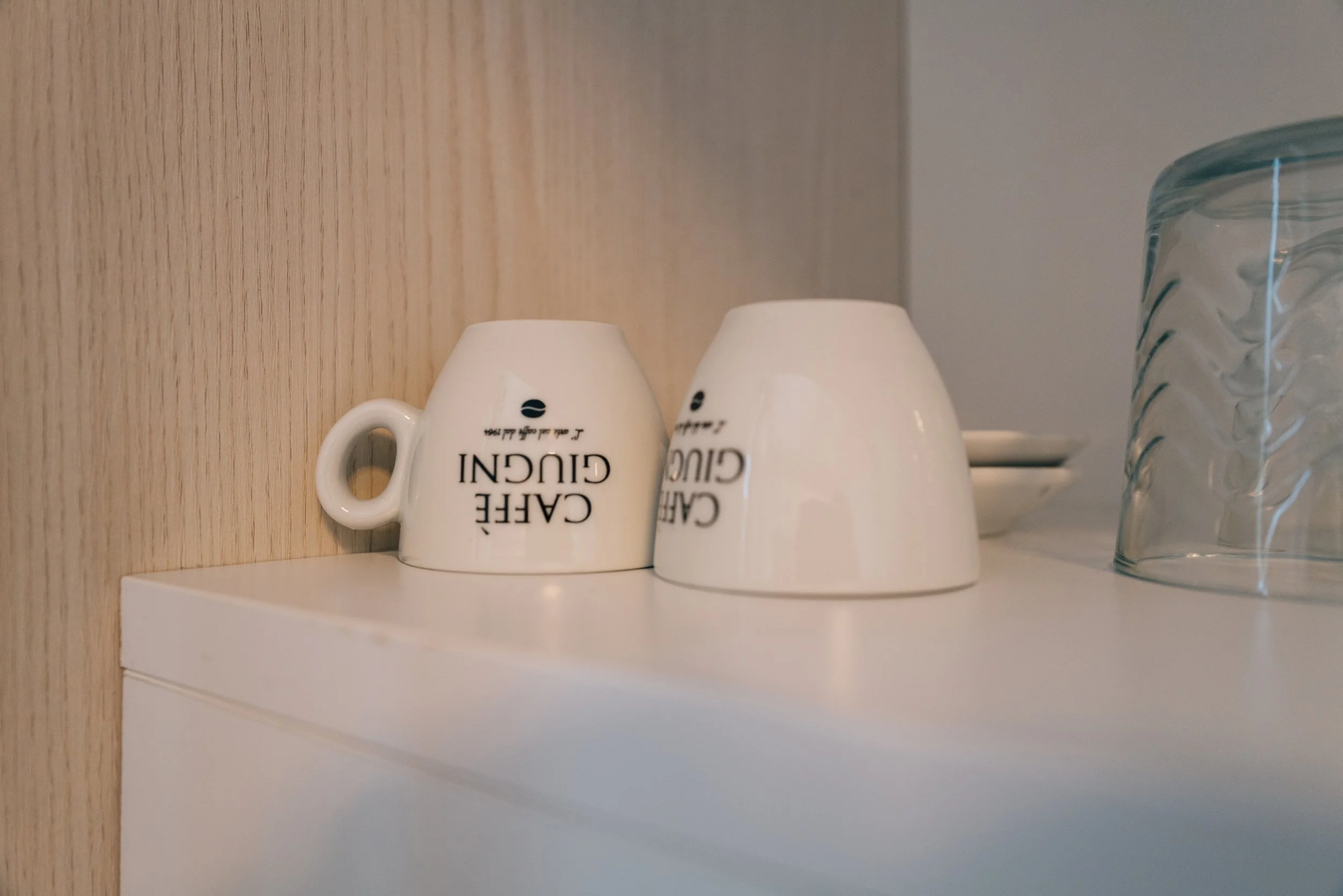 Due tazze da caffè con scritta 'Caffè GBCU' capovolte, una con manico, poste su una superficie bianca accanto a un contenitore di vetro con motivo ondulato.