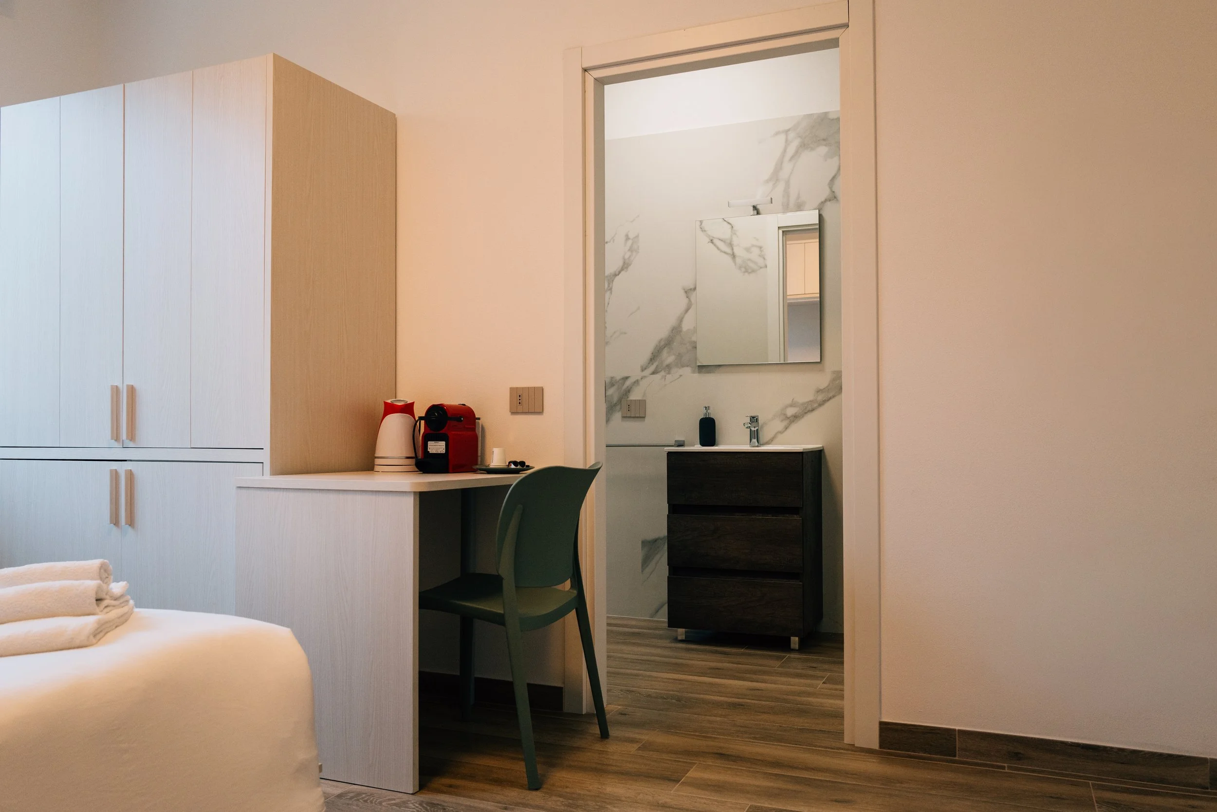 Angolo di una stanza con armadio, scrivania, sedia, e vista sull'area bagno con lavabo e specchio.