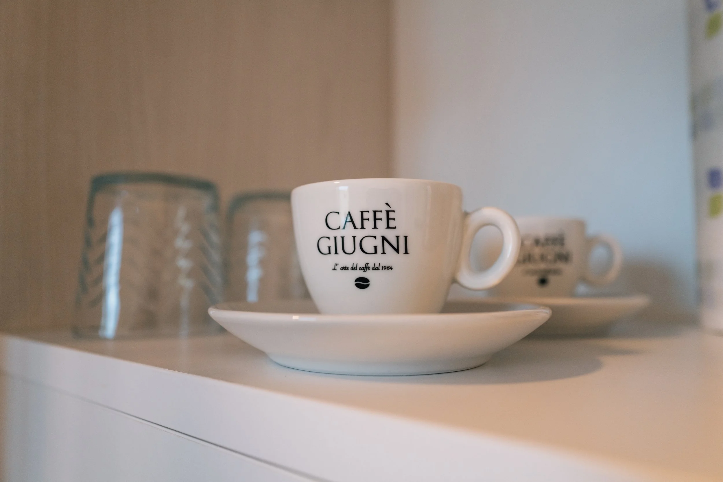 Tazza di ceramica bianca con scritta nera 'Caffè Giugni' e una tazzina uguale sul piattino, con tre bicchieri di vetro trasparenti e un altro piattino con un'altra tazza in sfondo.