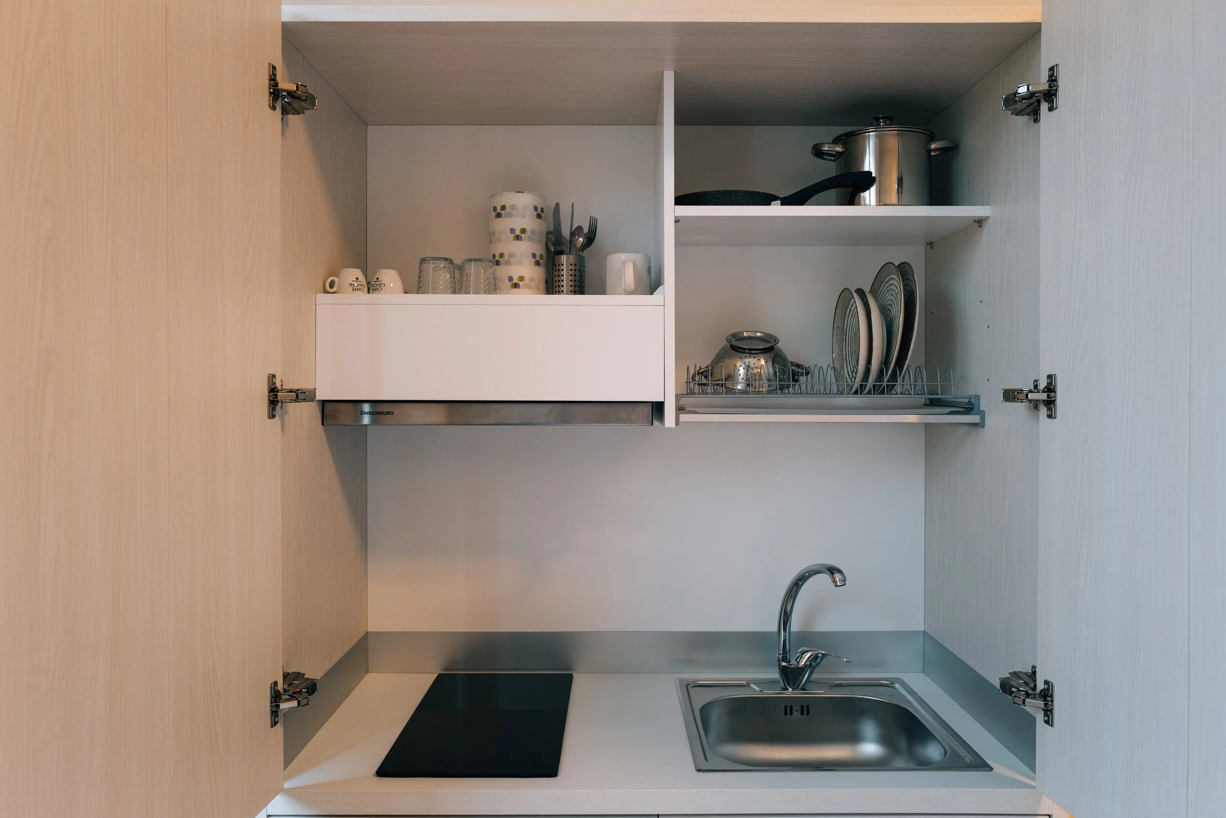 Armadio da cucina aperto con piatti, bicchieri e pentole, piano cottura e lavello.