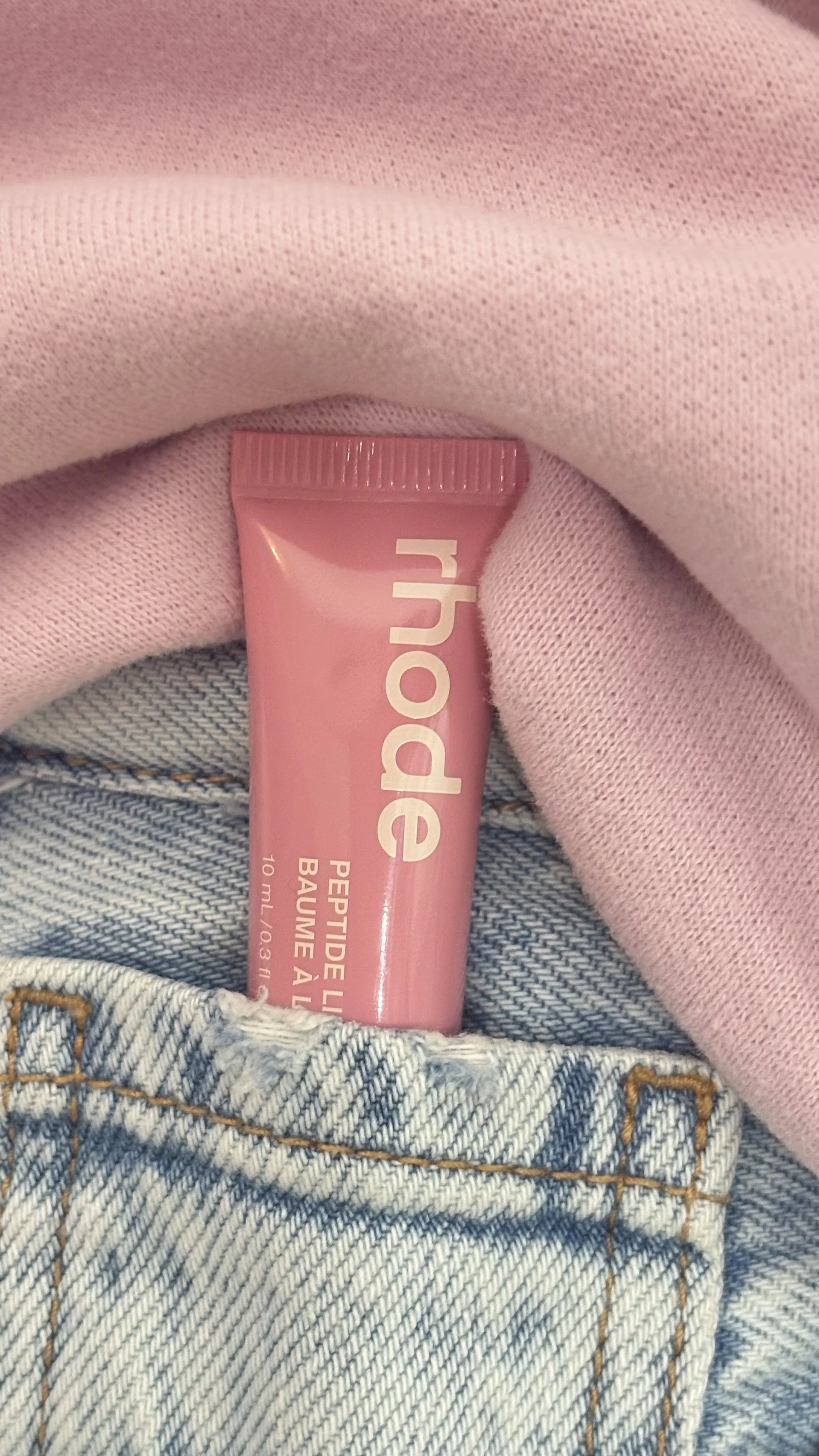 Pink tube of lip balm labeled 'huda' peeking out of a front pocket of blue denim jeans with orange stitching, with pink fabric waistband above.