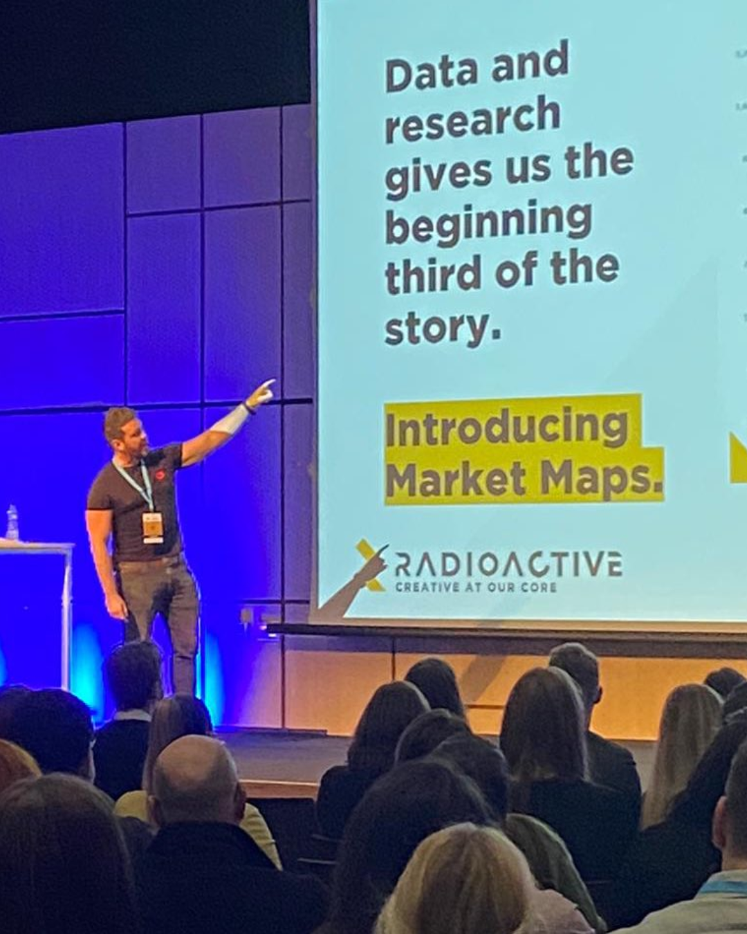 PR agency founder Rich Leigh giving a keynote speech. Leigh points at a large screen displaying a slide with text about data, research, and market maps at a conference with an audience.