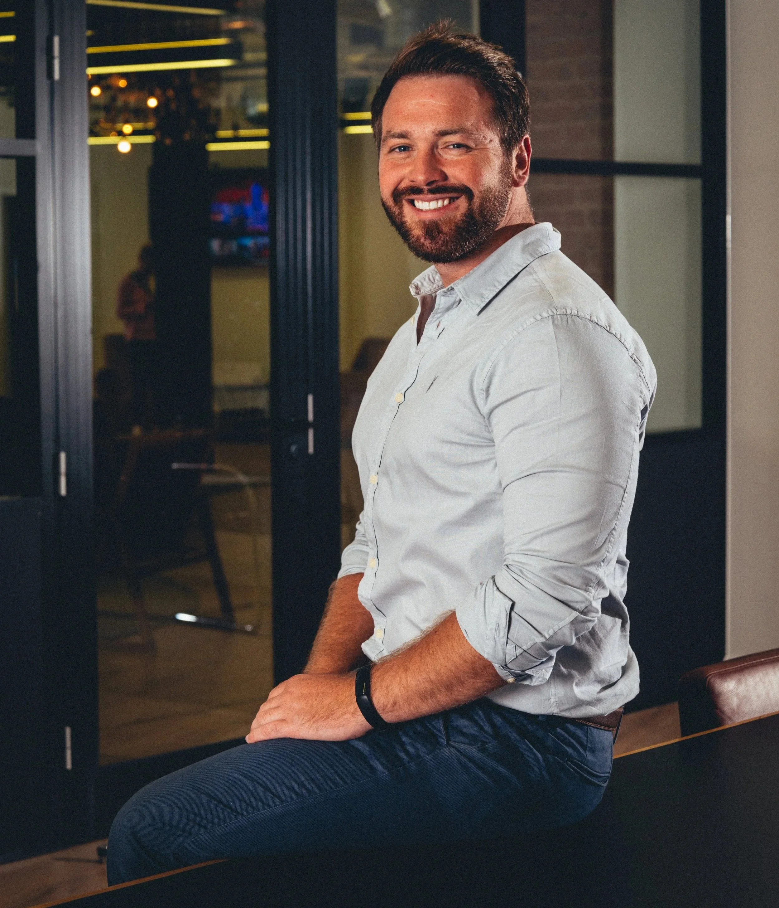 Rich Leigh in a light grey shirt and dark jeans sitting on a bench in a modern office or lounge area.