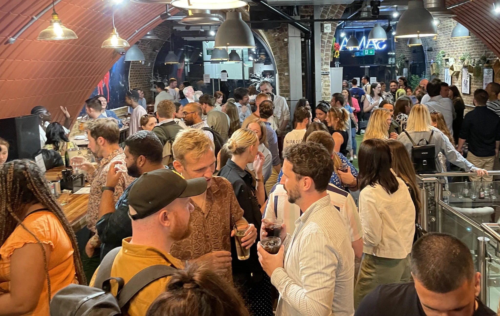 Crowded bar or restaurant filled with diverse people socializing and drinking in a lively, modern setting with brick walls, arched ceilings, and warm lighting.