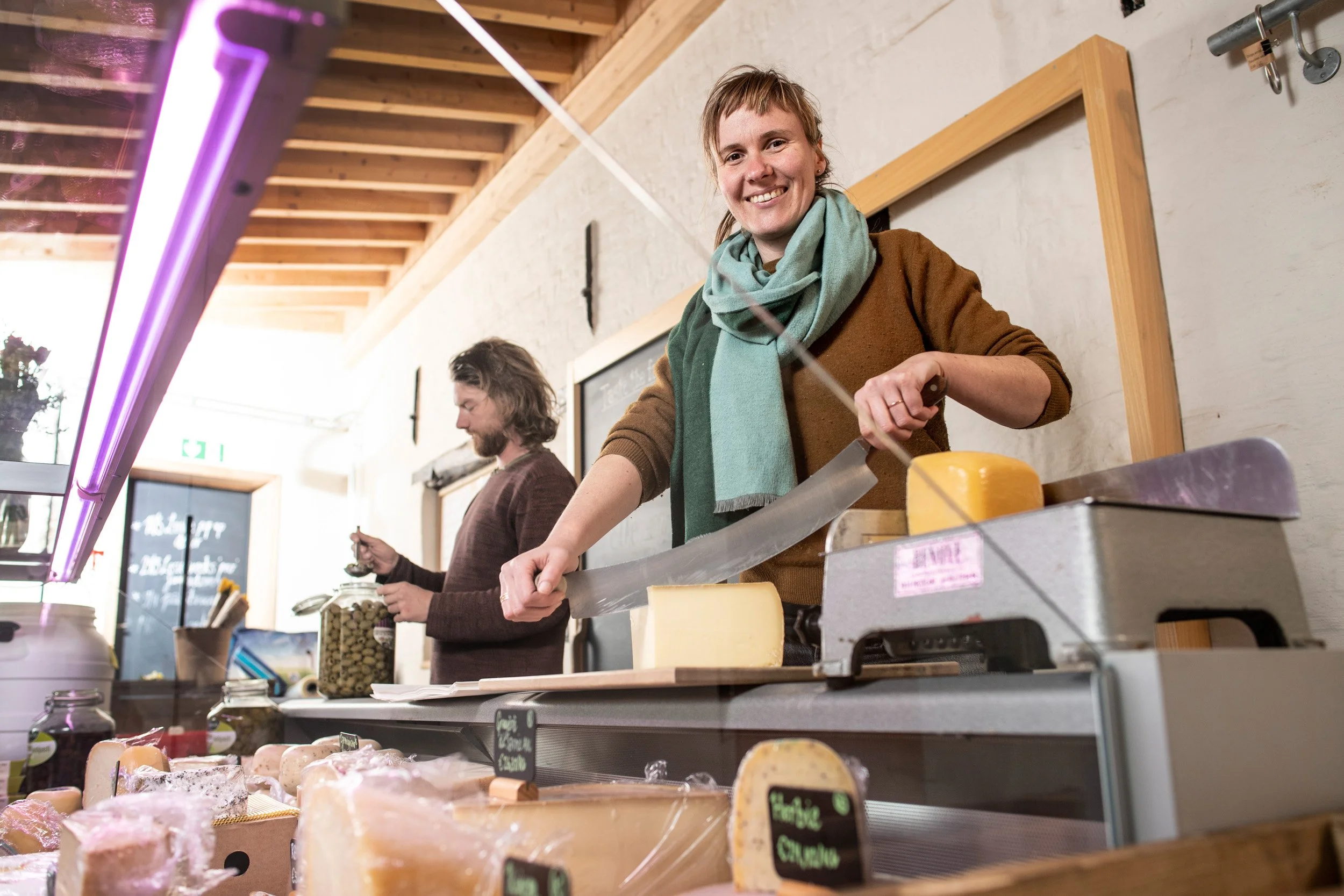 Een vrouw snijdt kaas in een kaaswinkel met andere klanten op de achtergrond.