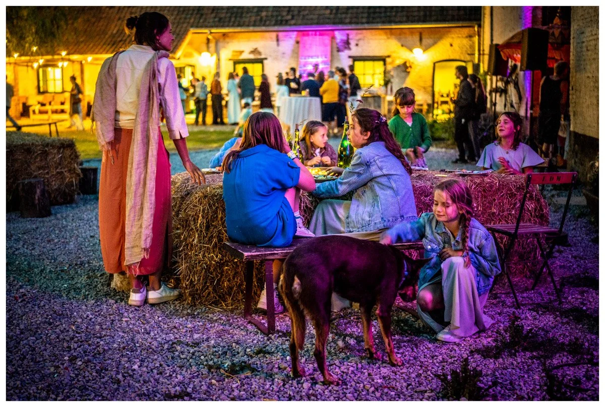 Kinderen en volwassenen zitten en staan rond een tafel met hooibalen tijdens een buitenfeest bij avond met kleurrijk verlichte achtergrond.