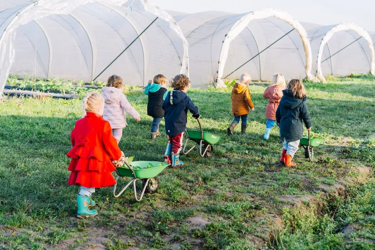 Kinderen met tuingereedschap die in een moestuin werken buiten, met kasstructuren op de achtergrond.
