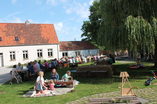 Mensen verzamelen zich op ons grasveld met picknicks en zitplaatsen, naast een grote boom en een rij huizen met rood dak, onder een blauwe hemel.