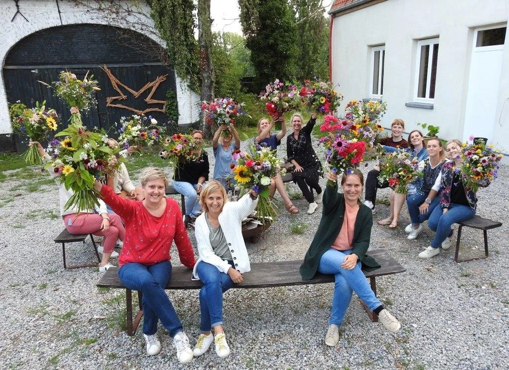Vijftien vrouwen die bloemen vasthouden en lachen in een tuin met houten banken en een witte muur.