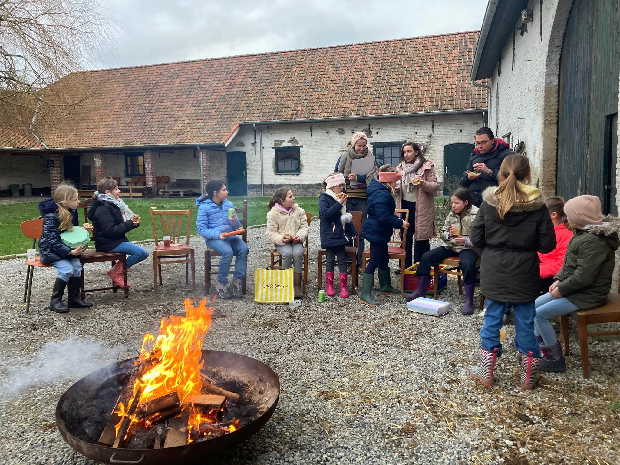 Een groep kinderen en volwassenen zitten buiten rond een kampvuur voor een oude boerderij met witte bakstenen muren en een rood dak. Het is een grijze dag en de kinderen lijken te genieten van snacks en drinken.