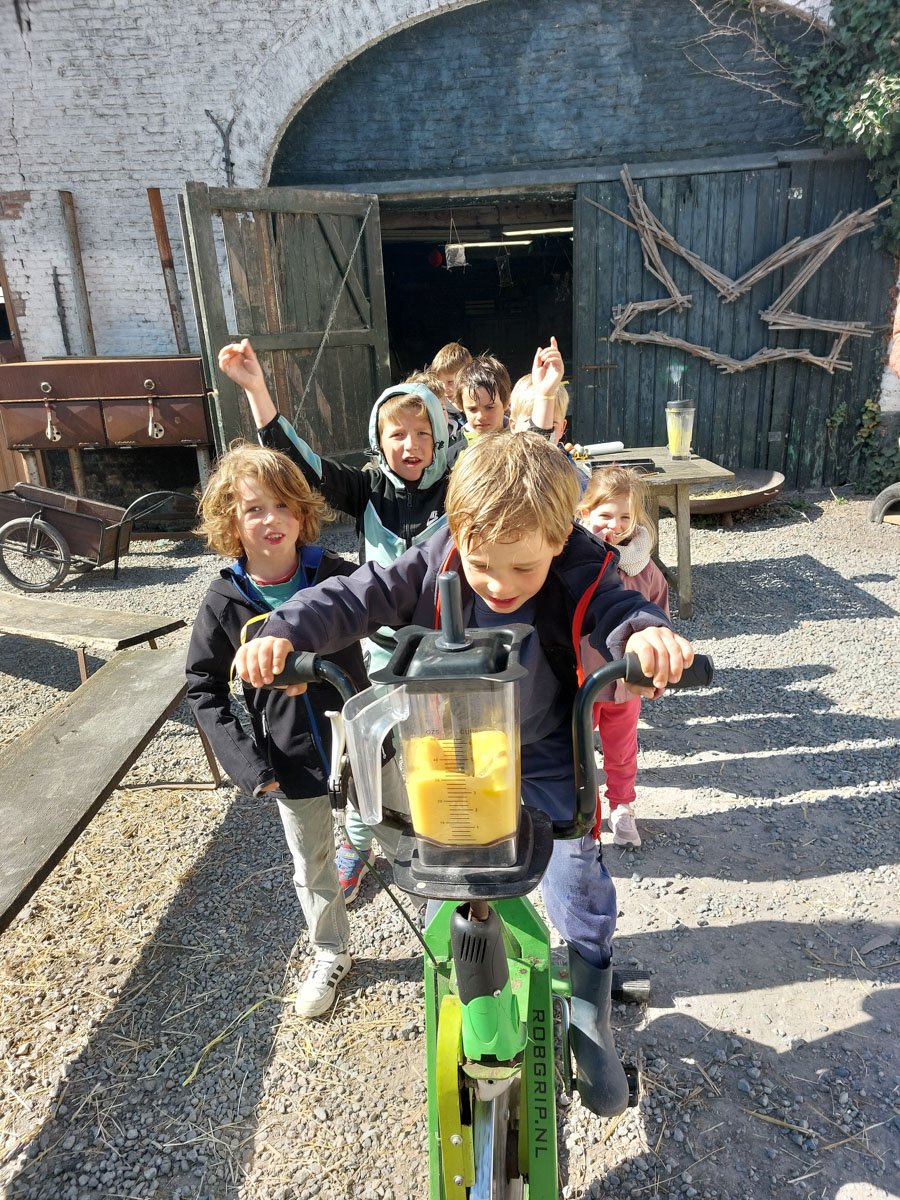 Kinderen spelen buiten, een jongen fietst op een groene pedallo met een blender die vol zit met een gele substantie en andere kinderen staan erachter en lachen.