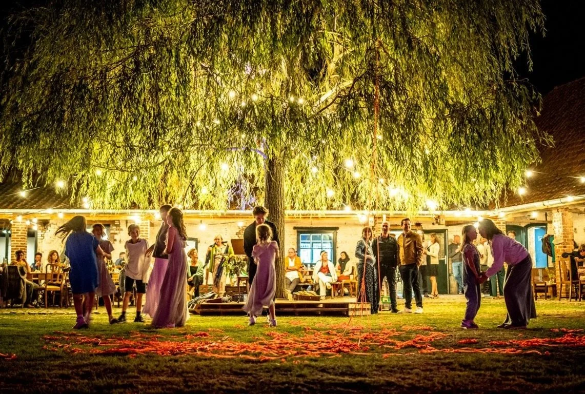 Gezellig buitenfeest met dansende kinderen onder een grote boom met lichtslingers, omringd door volwassen gasten bij een feesttent.