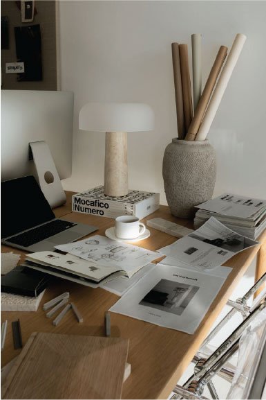 Desk with a lamp, a cup, open magazines, a closed laptop, and a large vase filled with rolled papers.