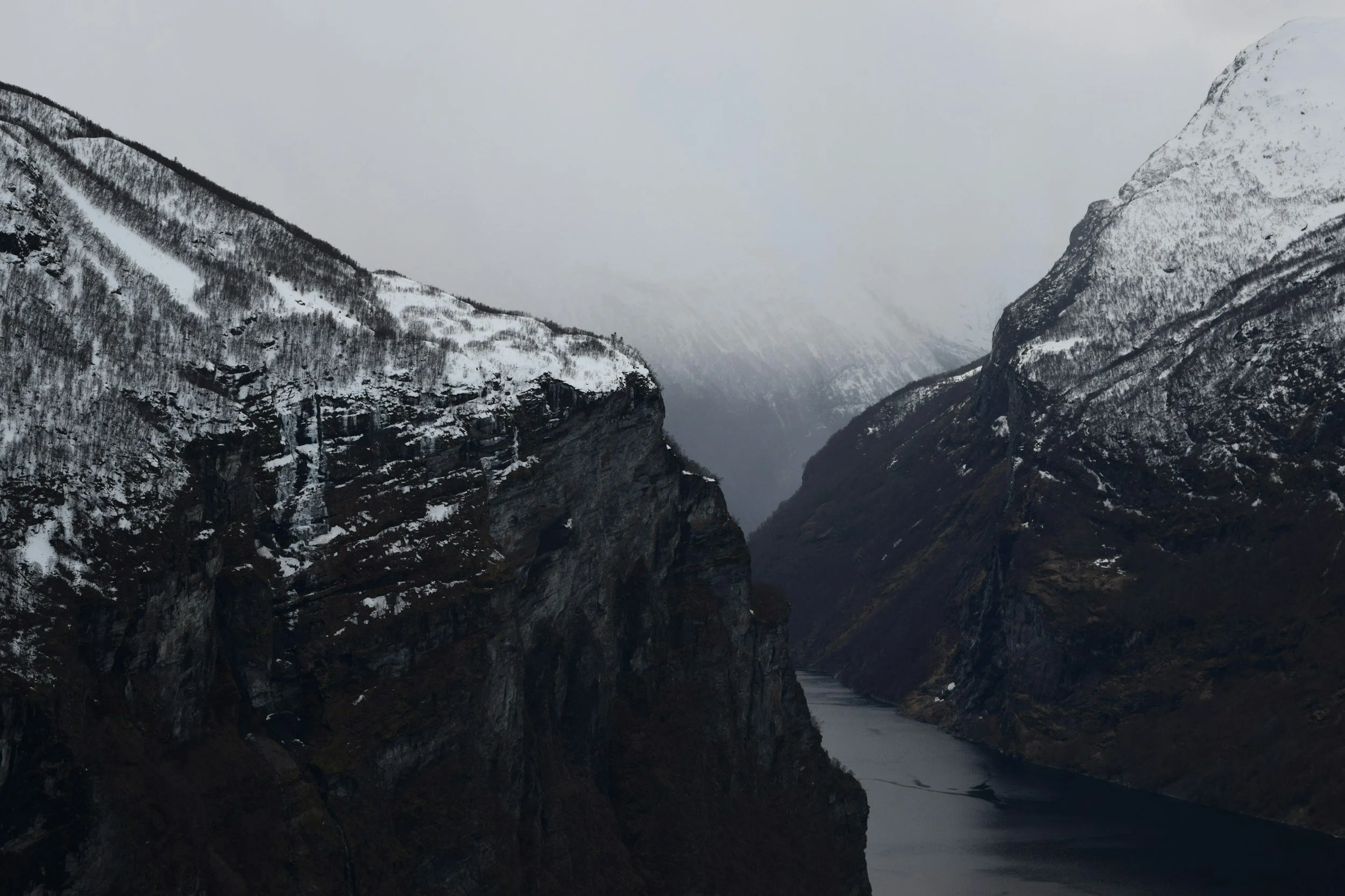 Snow-covered mountains with a river running between steep cliffs, foggy and overcast sky.