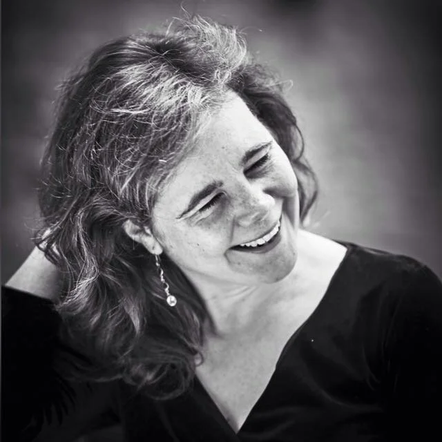 A woman with wavy hair smiling and looking to the side in a black top, in a black-and-white photo.