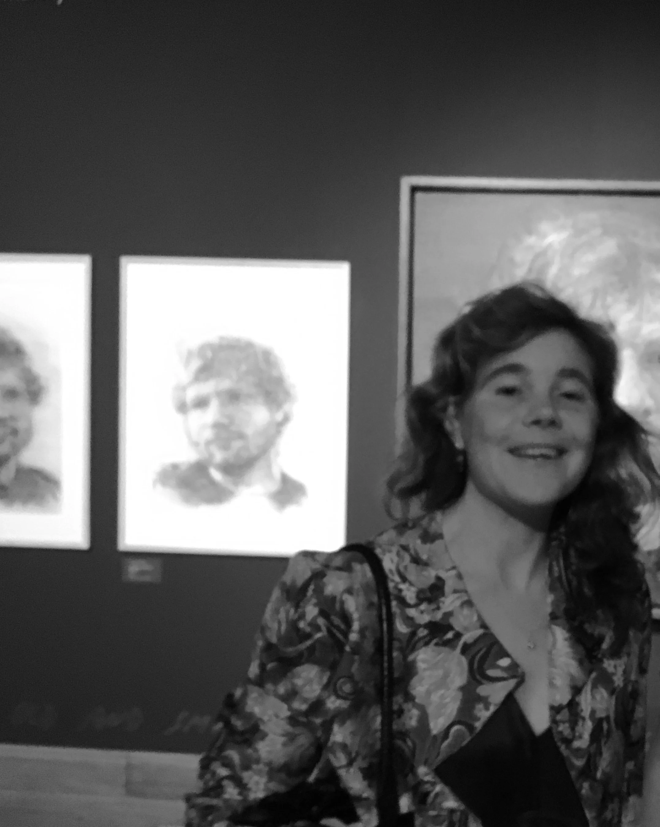 A woman with curly hair, wearing a floral-patterned top, smiling, standing in front of two framed black-and-white photographs on a dark wall.