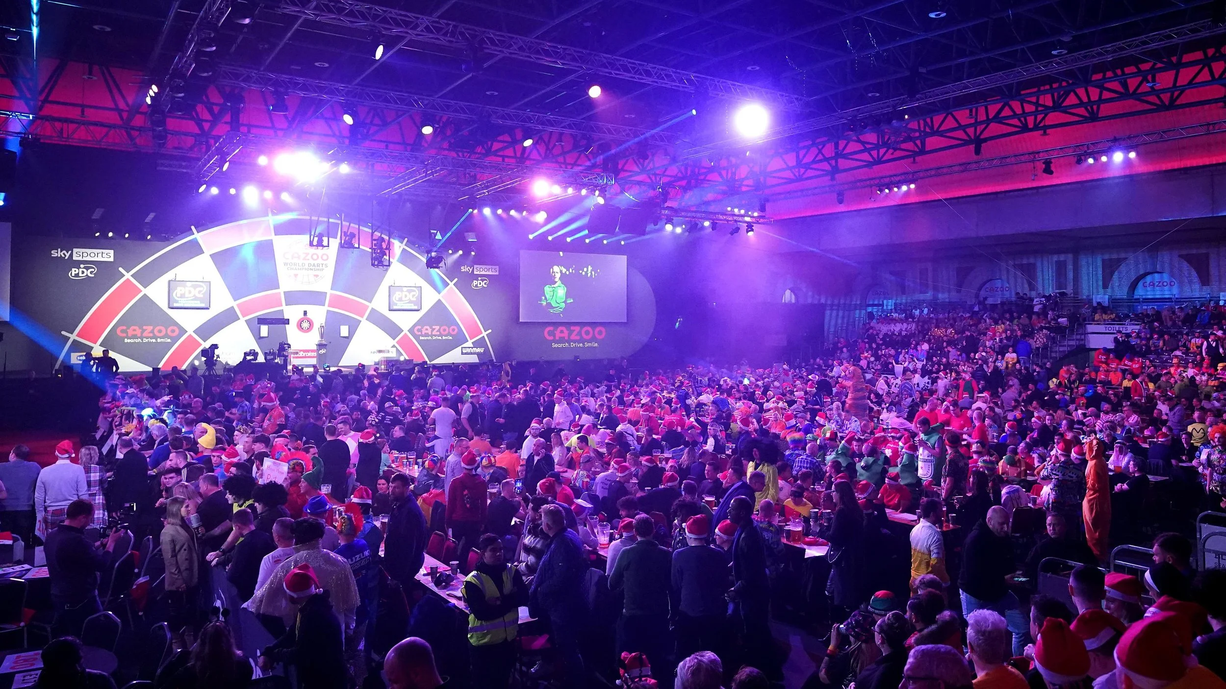 Indoor arena filled with a large audience, many wearing Santa hats, watching a darts competition on a brightly lit stage with multiple screens.