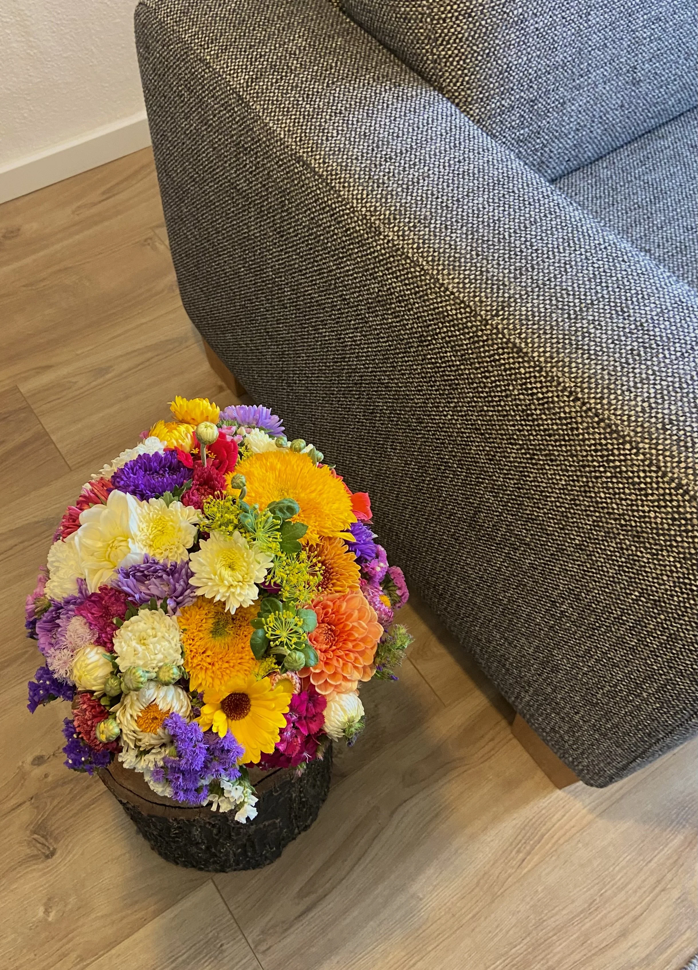 Bunter Blumenstrauß neben dunklem Sofa auf Holzboden.