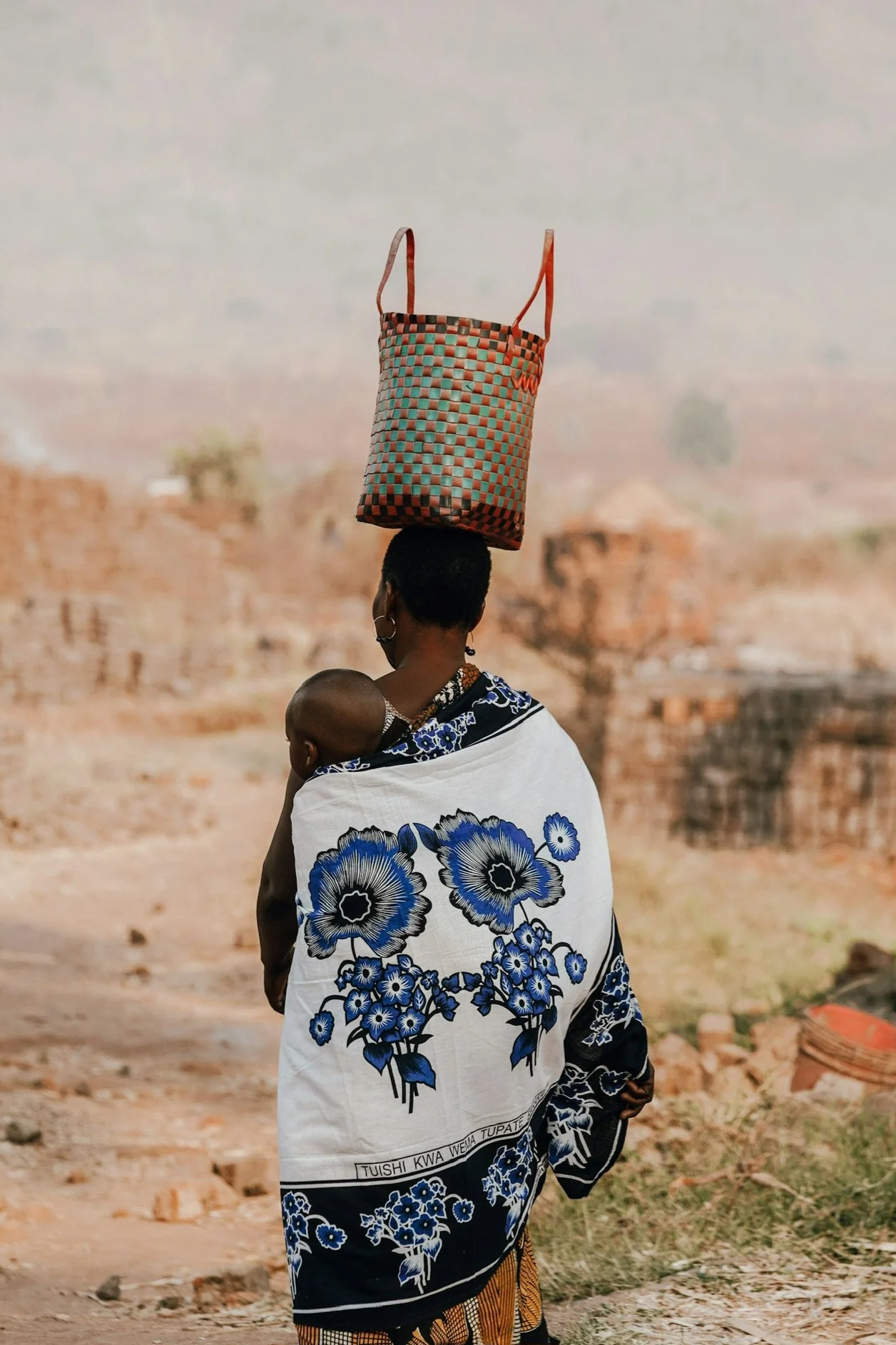 A woman carrying a child on her back, wrapped in a white and blue floral shawl, with a woven bag balanced on her head, walking through a rural area with ruins and a distant landscape.