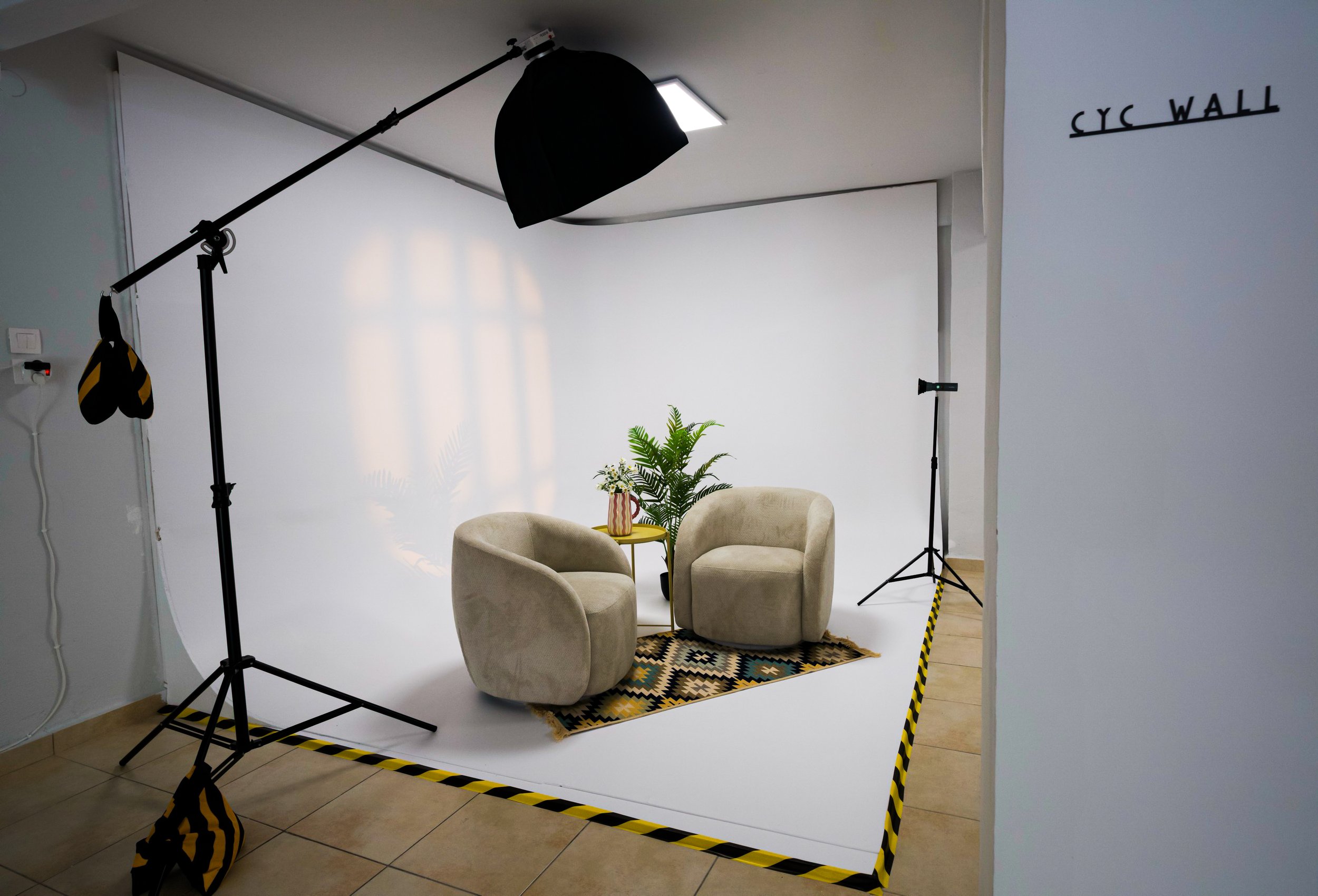 A photography setup with studio lighting, a white backdrop, and furniture including a chair and a small table with a potted plant, labeled 'CYC WALL' on the wall.