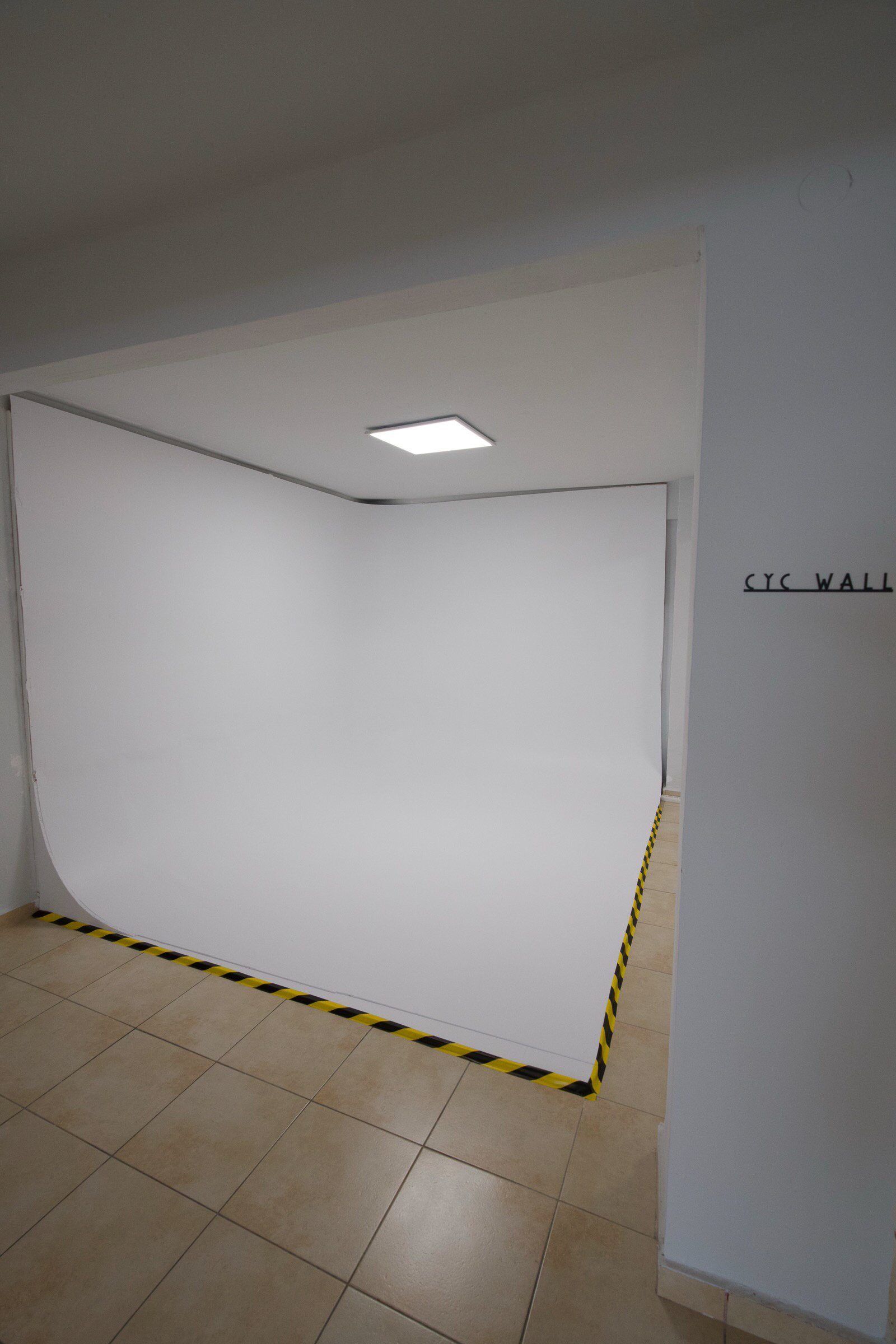 A white photography backdrop set up inside a room with tiled floor, surrounded by black and yellow caution tape. There is a ceiling light above the backdrop, and a partial sign on the right wall that reads 'CYL WALL'.