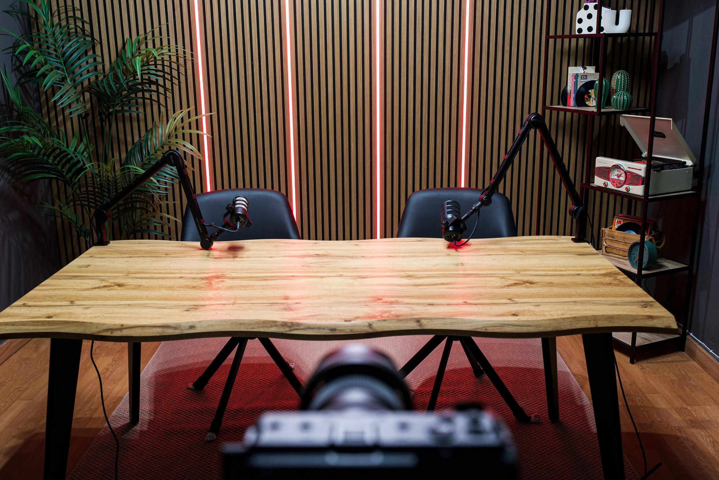 A podcast recording setup with two microphones on adjustable arms, a wooden table, black chairs, a potted plant on the left, a decorative bookshelf on the right, and a wooden wall with vertical red LED lights in the background.