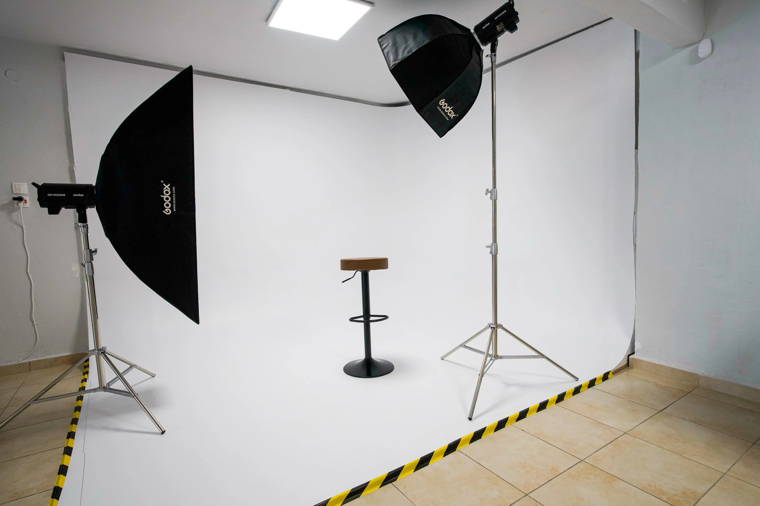 Photography studio setup with a white backdrop, two light stands with softboxes, a stool in front of the backdrop, and a yellow and black caution tape marking the floor.