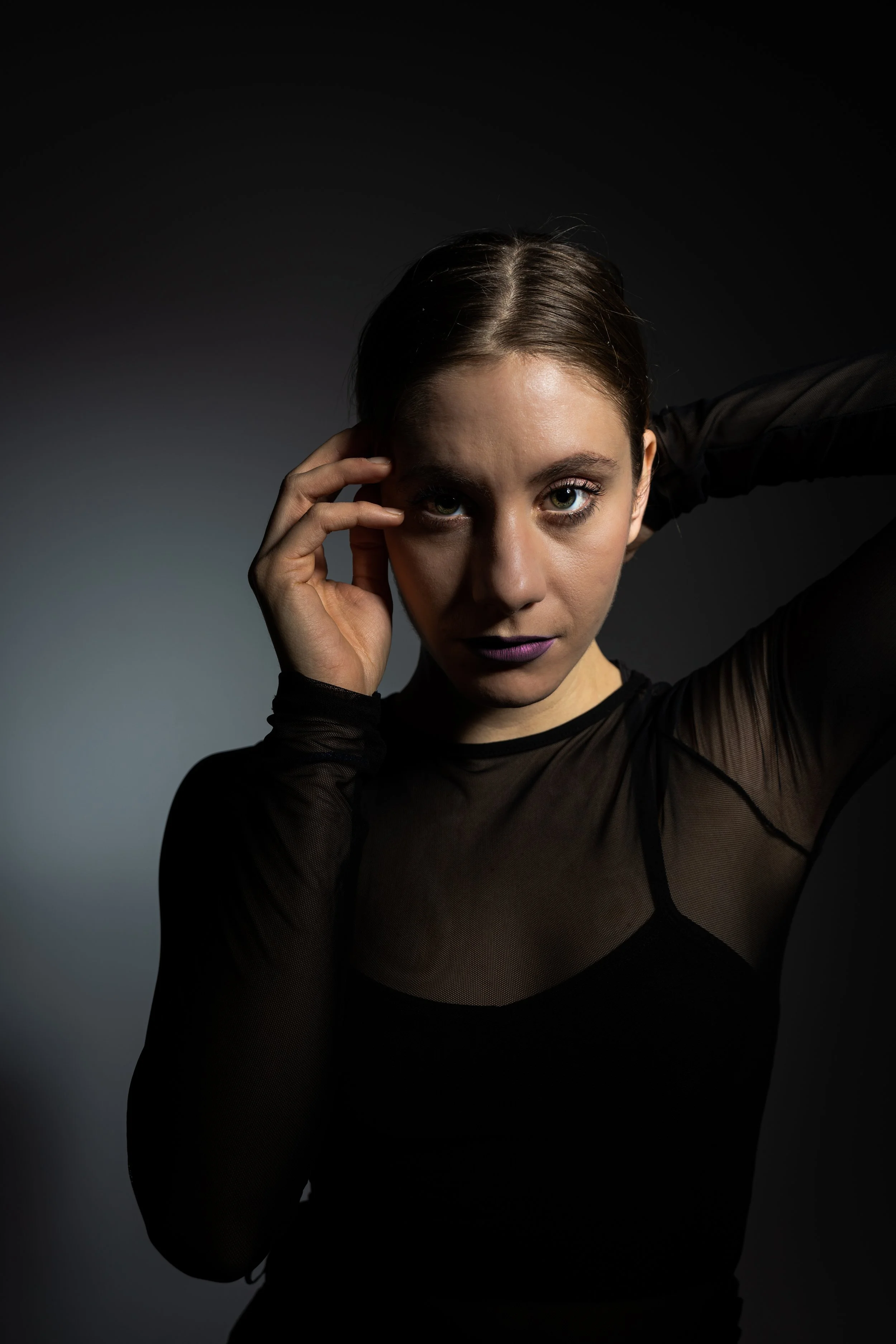 A woman with dark hair and striking eyes poses against a dark background, wearing a sheer black top with intricate detailing, and touches her temple with her left hand.