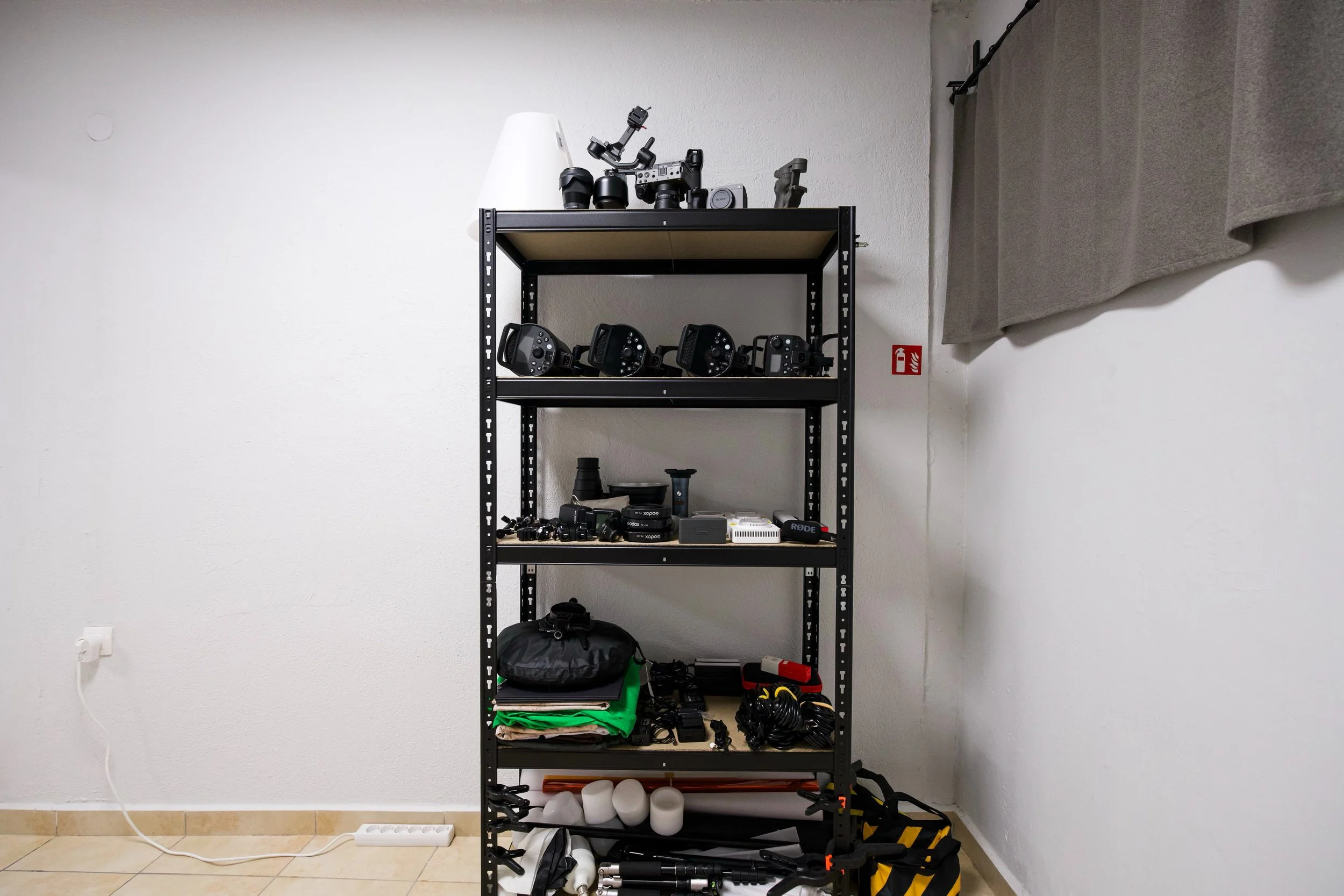 A black metal shelf with four tiered levels, holding various photography and videography equipment. The top shelf has cameras, camera lenses, and a light. The second shelf has multiple camera triggers or remote controls. The third shelf has a black bag, electronic devices, and neatly folded green and beige cloths. The bottom shelf contains rolled-up white papers or foam, tripod stands, and other gear. The shelf is against a white wall, with a gray curtain draped on the right side and a power strip plugged into a wall outlet on the left.
