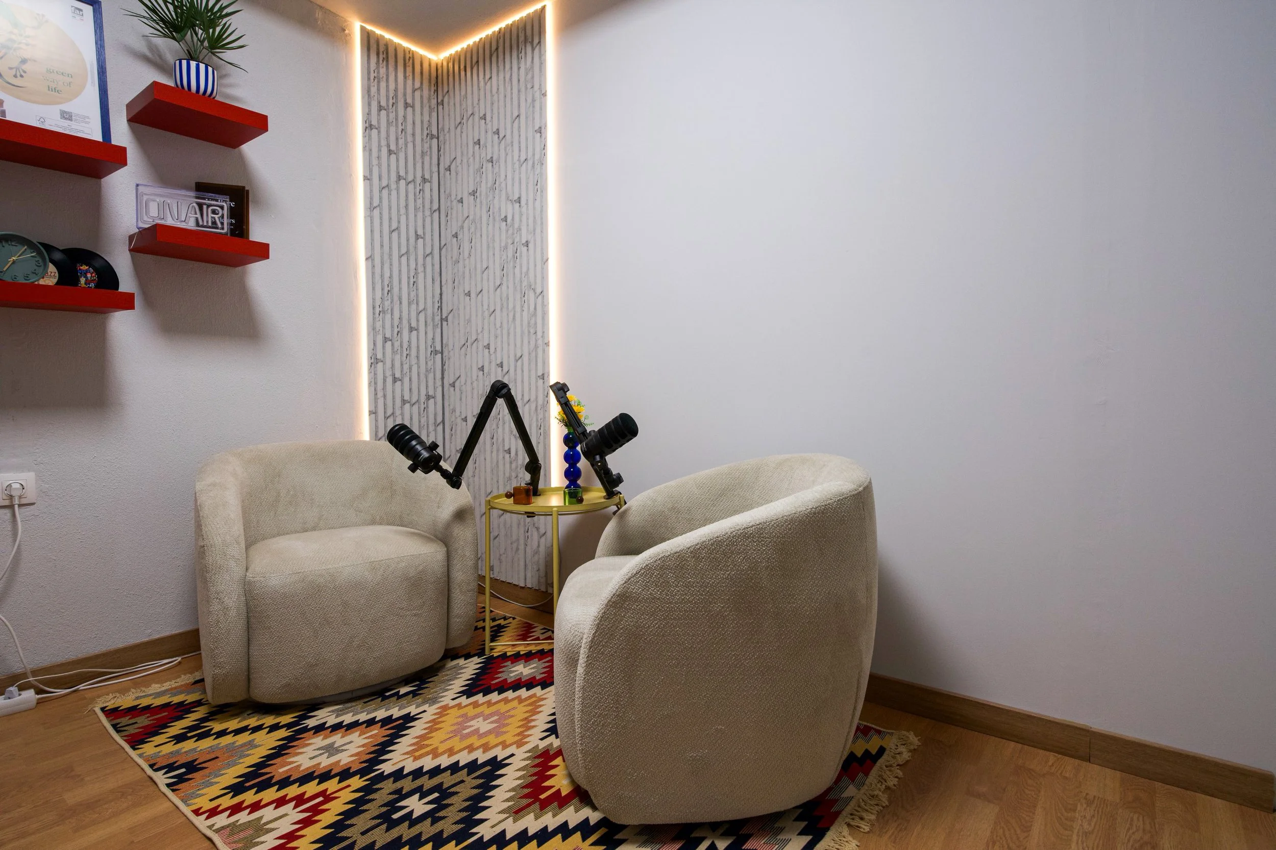 A cozy sitting area with two beige armchairs placed around a small yellow side table holding a camera and a colorful decorative item. There is a patterned area rug on a wooden floor, a textured wall with vertical white and gray stripes, and wall-mounted red shelves with decorative items including clocks, books, and a plant. The room features modern LED lighting along the ceiling and wall corners.