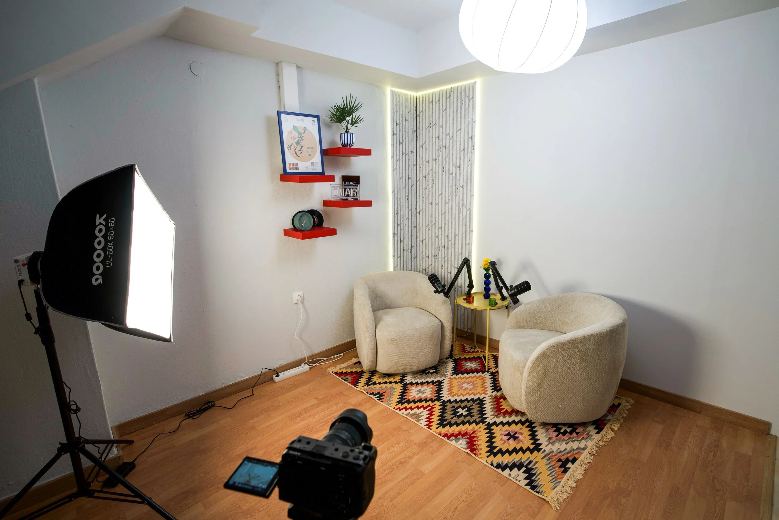 A cozy room setup with two beige armchairs, a colorful geometric rug, and a small yellow table with toys. There are three red wall shelves with decorations, including a framed picture, a potted plant, and a clock. A lighting panel is on the ceiling, and a photo or video camera is positioned in front of the scene, with professional lighting equipment to the side.