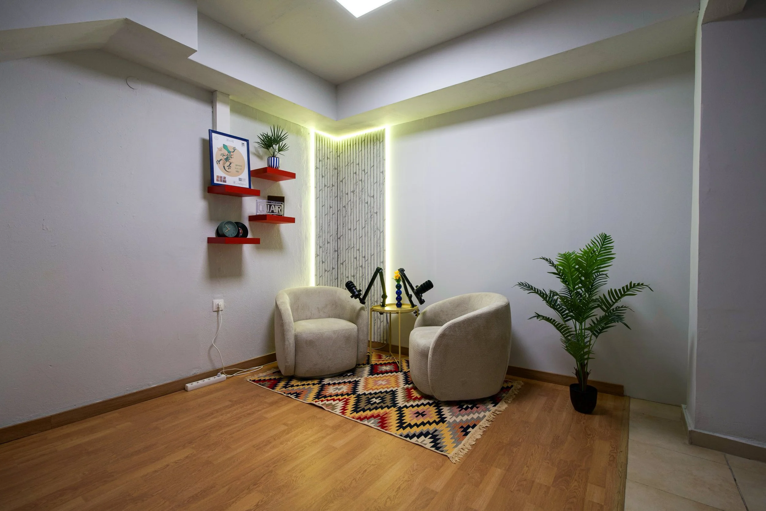 A cozy corner with two beige armchairs around a small round yellow table, a colorful geometric rug, potted plants, and wall shelves with decorative items in a room with white walls and ceiling lighting.