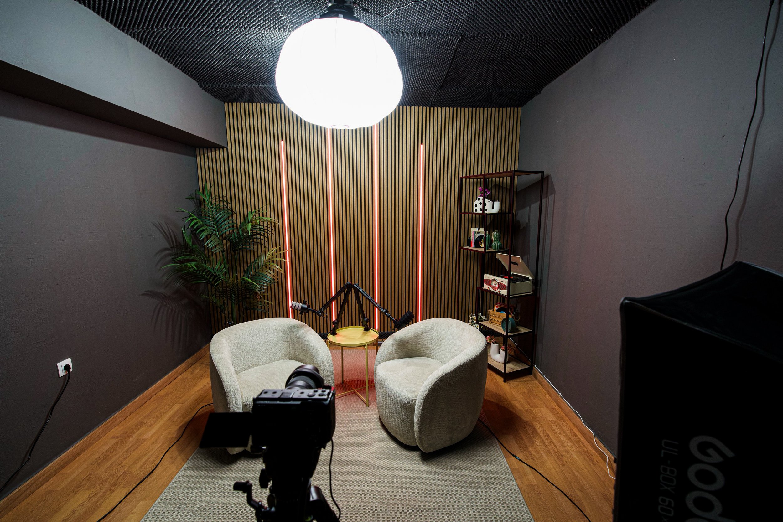 Interior of a room with two armchairs, a small round table, a tall black shelving unit with decorative items, a large round ceiling light, and a wooden slat wall with vertical pink lighting accents, all with dark gray walls and wooden flooring.