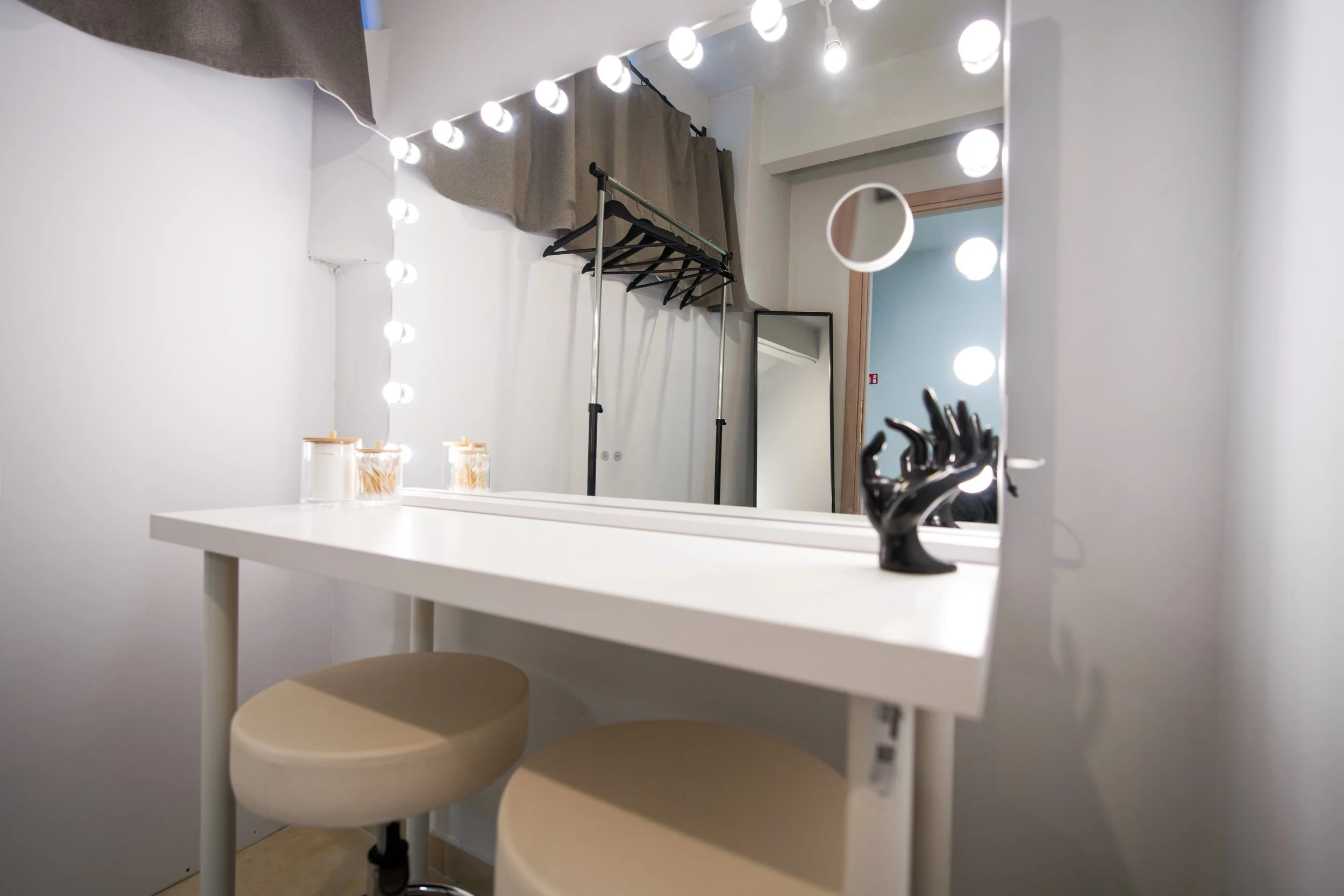 A white makeup or vanity table with a large mirror surrounded by round bright lights. In front of it, two beige stools are visible. On the table, there are three jars filled with what appears to be cotton swabs or similar items. In the background, a clothing rack with several black hangers and a gray curtain are visible, as well as part of a doorway and a mirror reflecting part of the room.