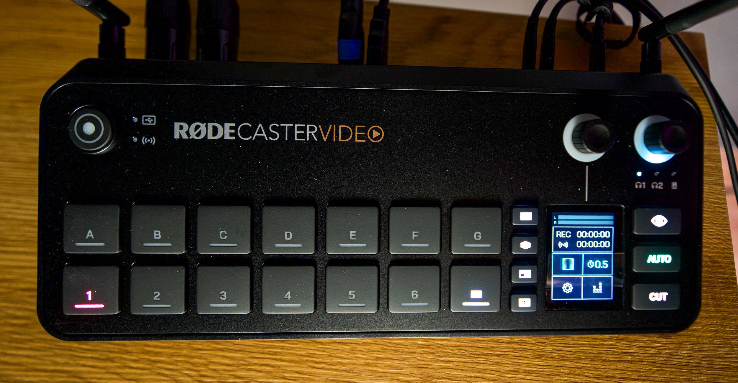 A black RODECaster Video device with a small screen displaying recording settings, connected with multiple cables, placed on a wooden surface.
