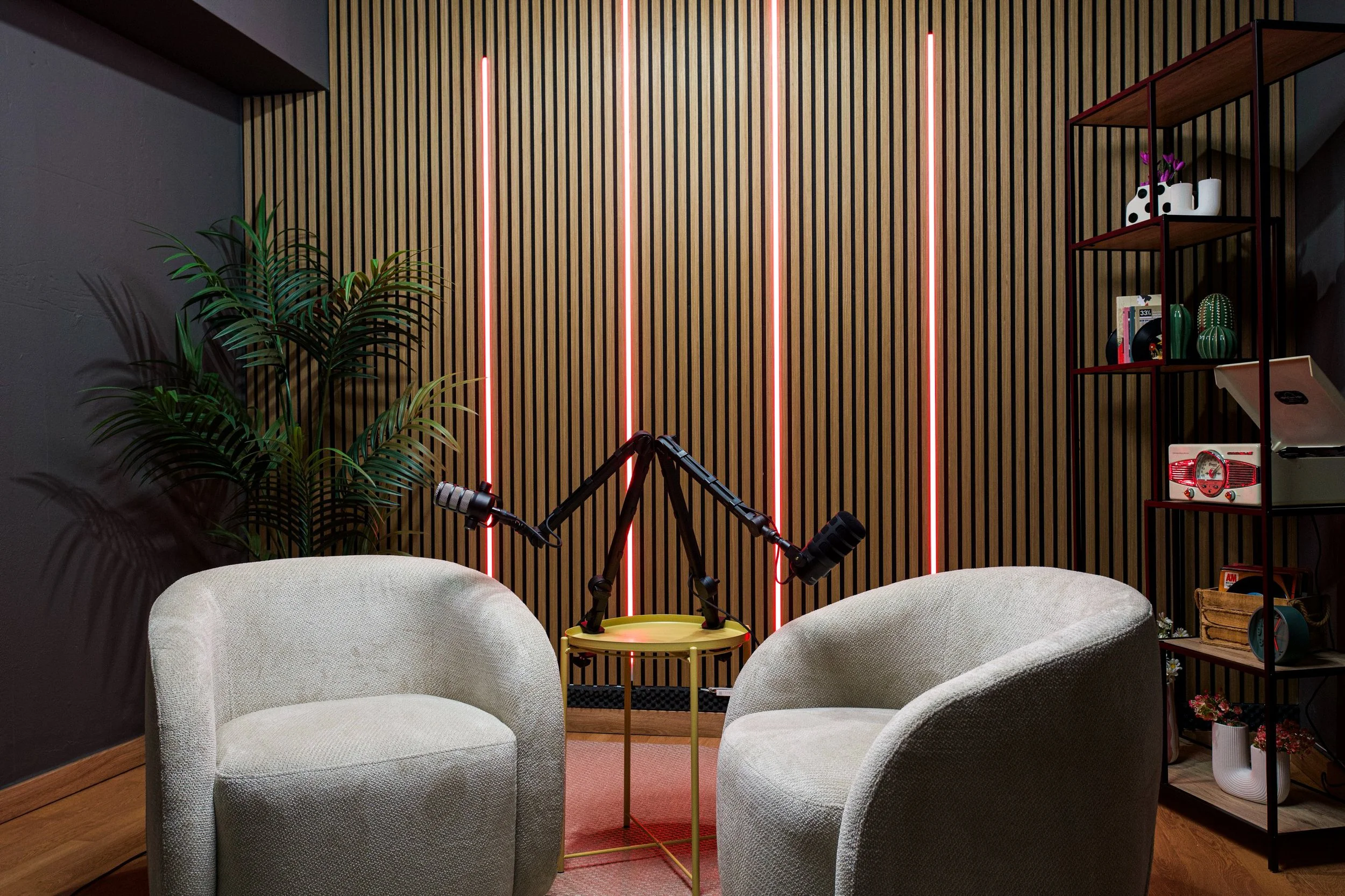 Interior living room with two white armchairs, a yellow side table with microphones, a large decorative wooden wall with pink LED lights, a black shelf, and a tall plant.