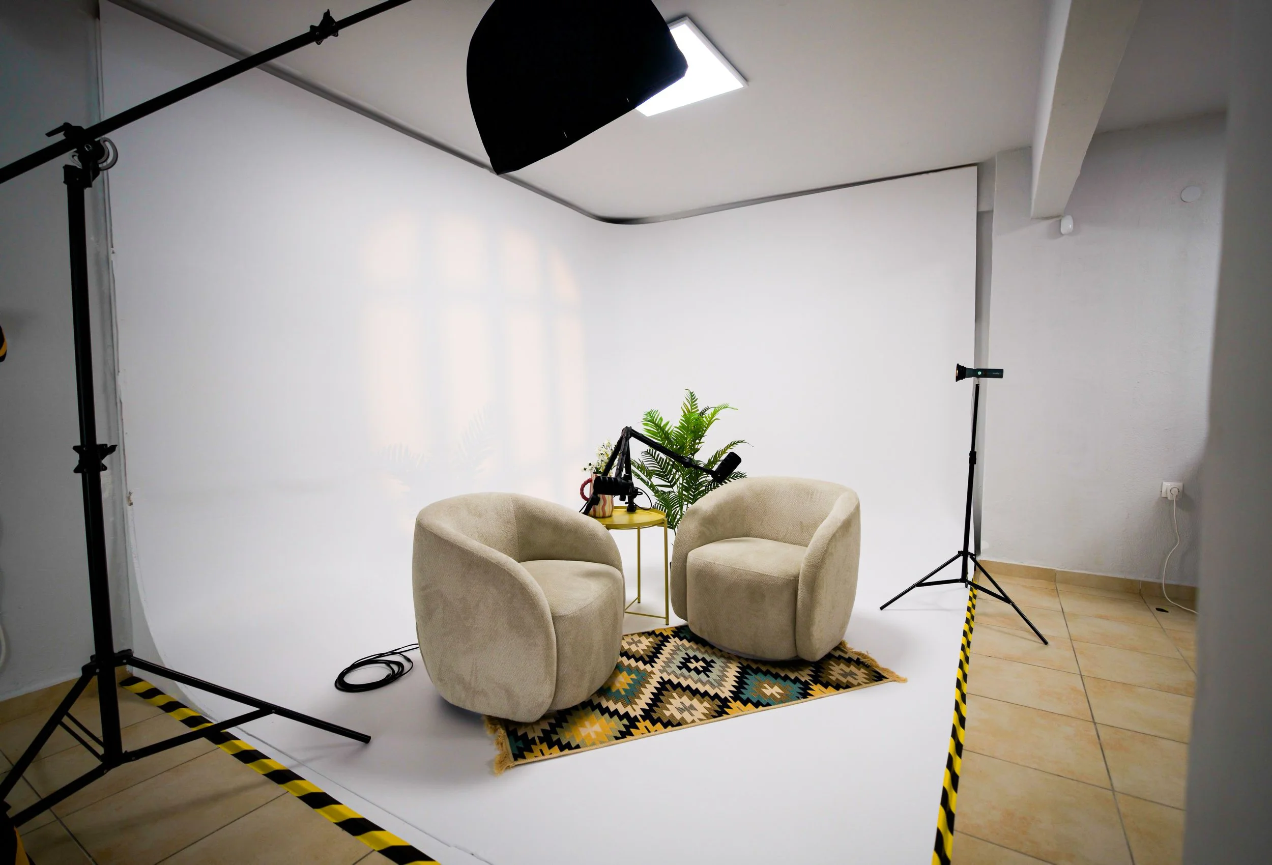 Professional photography studio setup with a white backdrop, two beige chairs, a small yellow table with potted plants, studio lights, and safety tape on the floor.