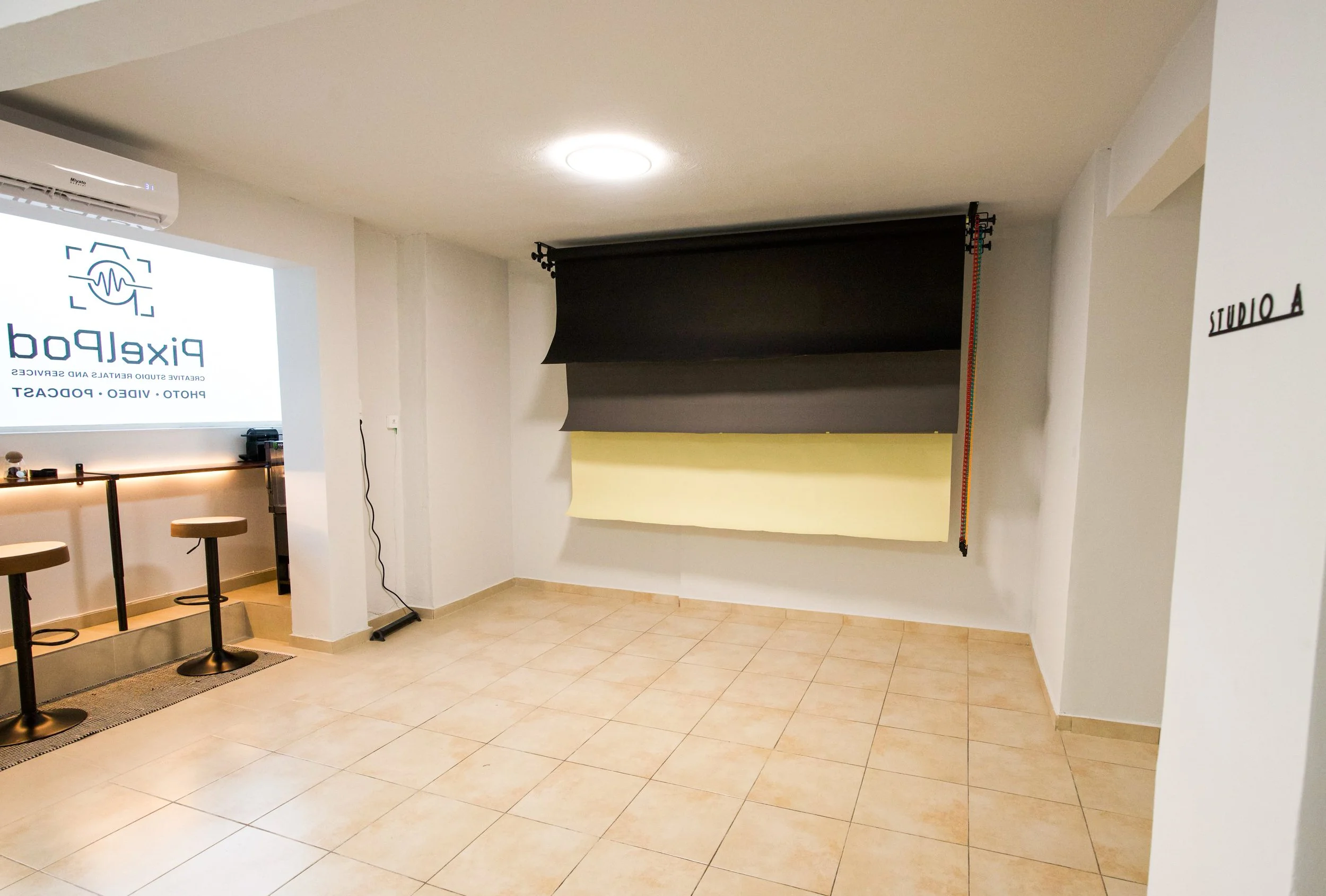 An empty room with white walls and beige tiled floor, featuring a photography backdrop with black and yellow paper, an air conditioning unit, and a small bar area with stools.